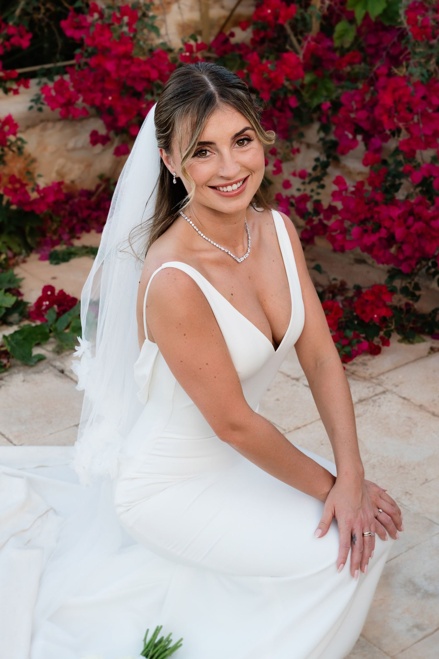A bride in a white wedding dress poses against a backdrop of vibrant red bougainvillea flowers on a stone patio.