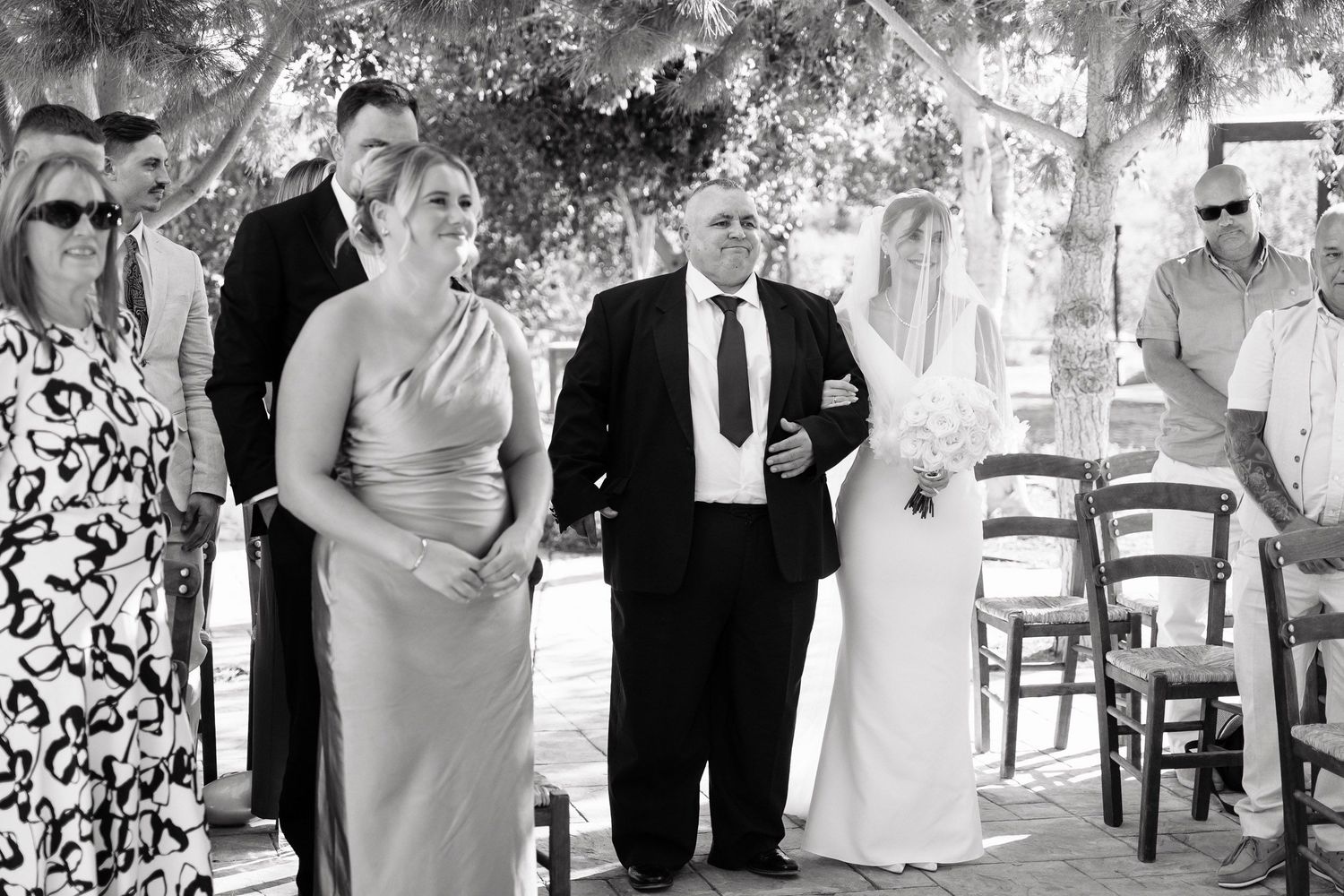 Black and white photo of a formal wedding gathering under a tree-covered outdoor venue with guests in formal attire.