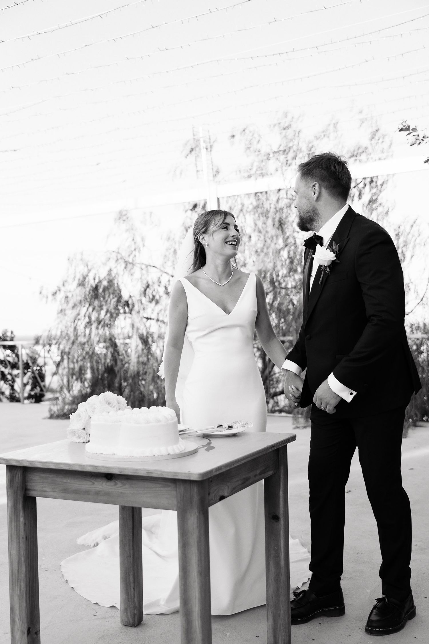 A wedding couple shares a playful moment at three wooden tables in an outdoor setting with white decor.