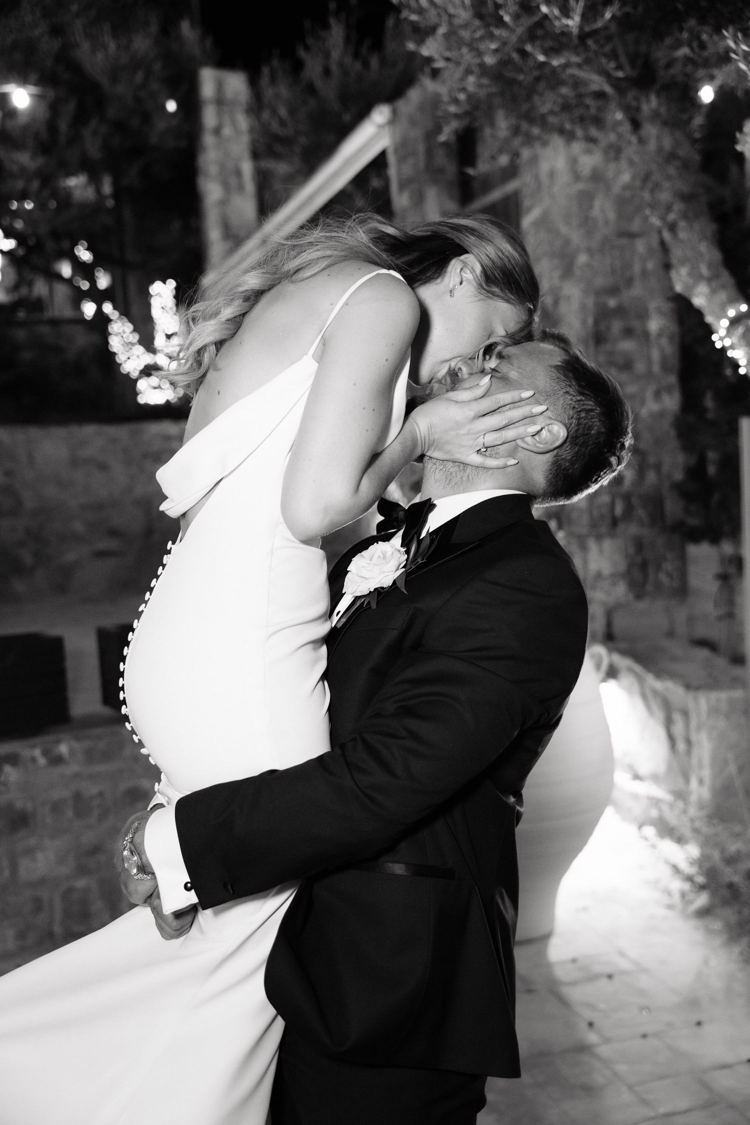 A romantic black and white wedding photo showing a couple sharing a passionate kiss outdoors at night with twinkling lights.