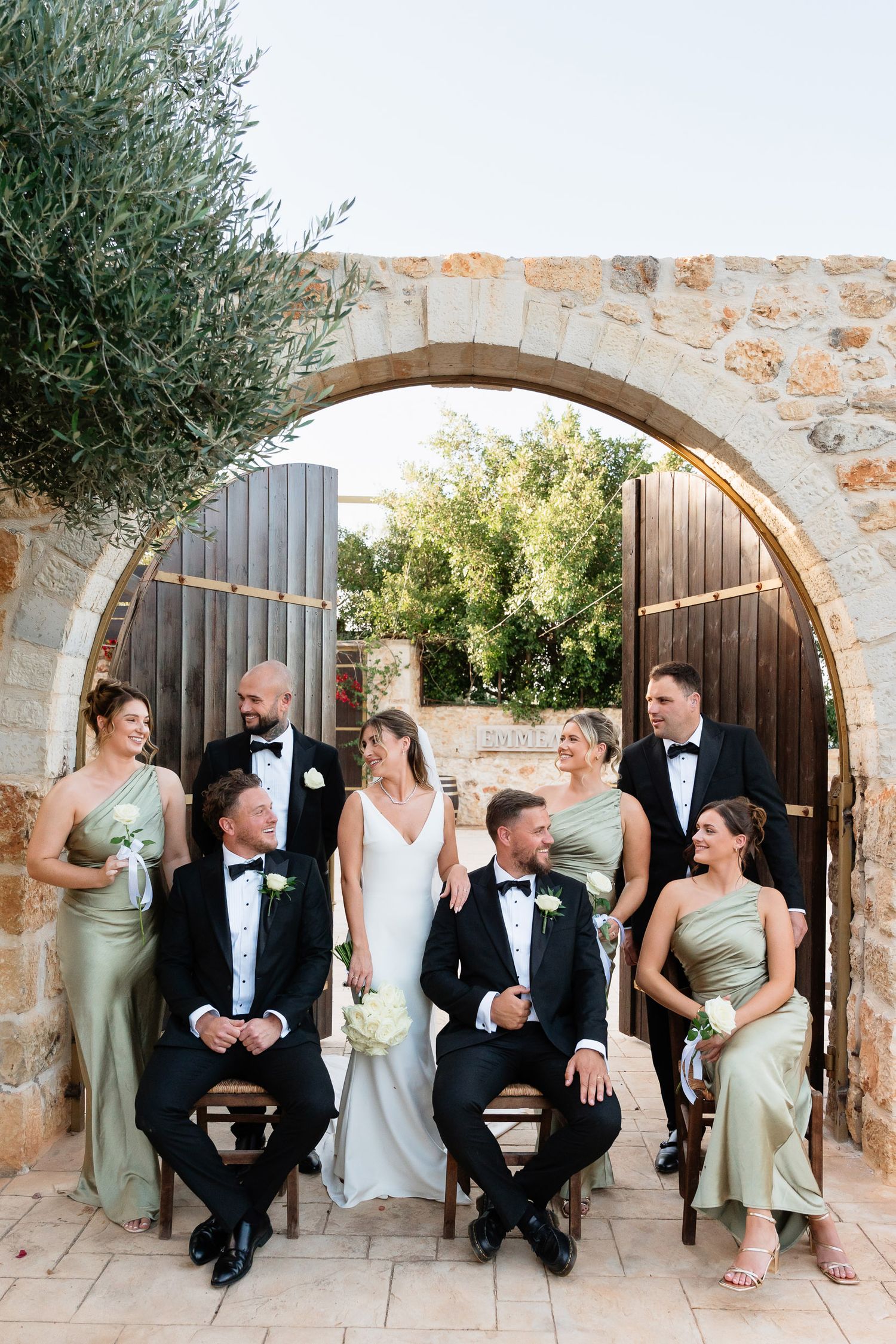 Wedding party poses together under a rustic stone archway with olive trees and wooden doors in the background.