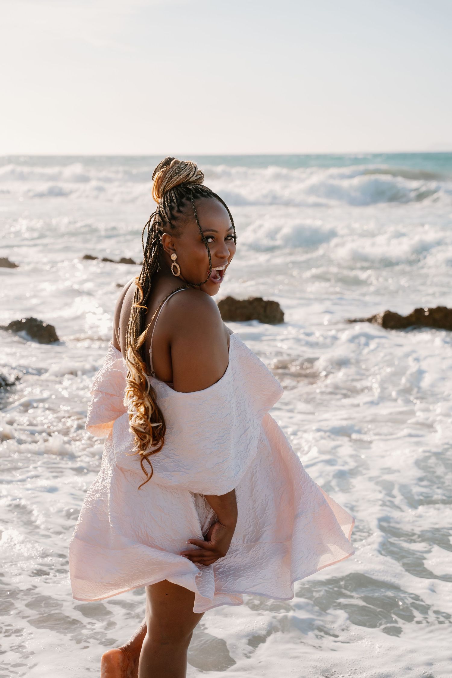 Woman in white dress twirls gracefully on a sunny beach with ocean waves crashing against rocks in the background.