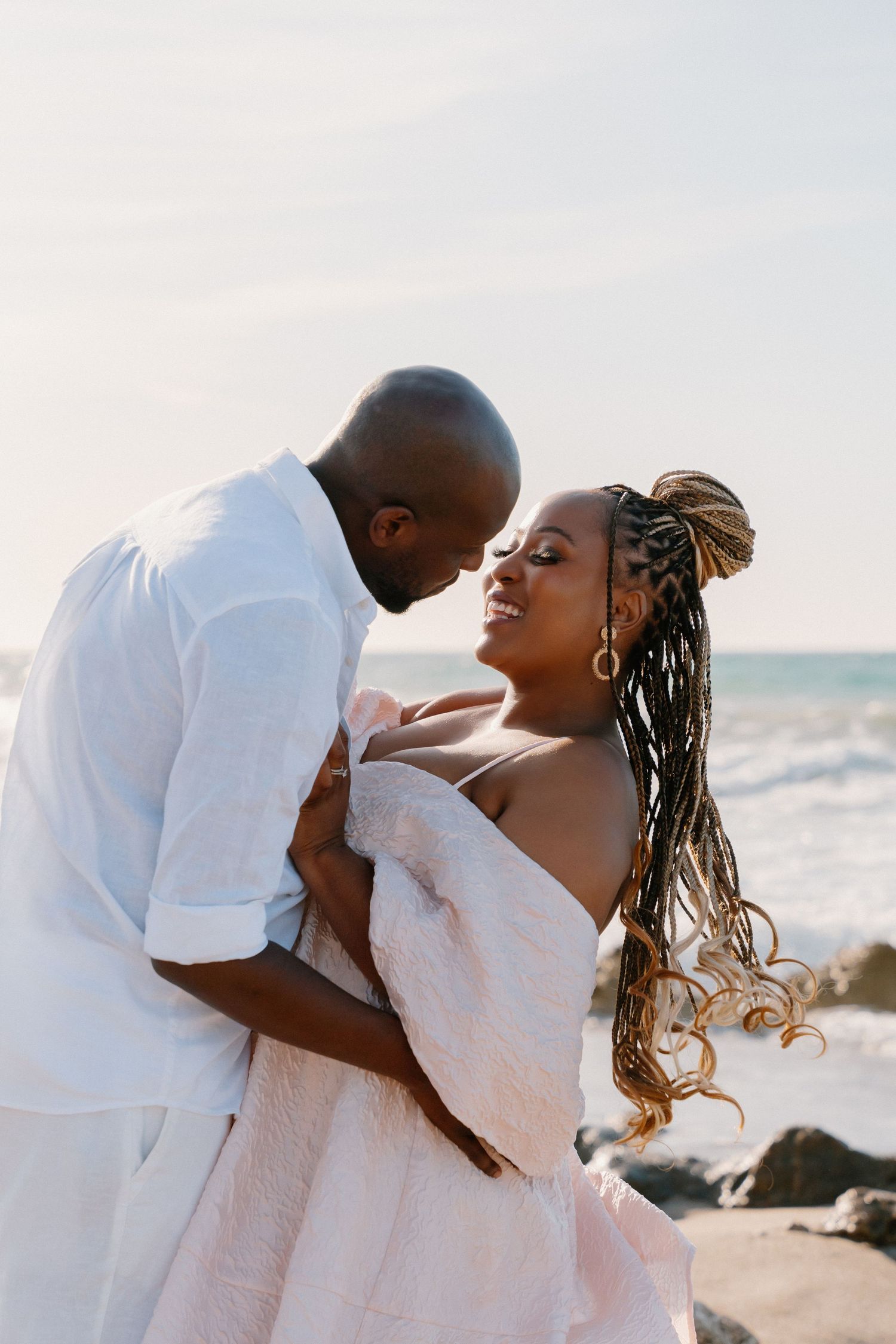 A romantic couple embraces and laughs together during a beach photoshoot at sunset.