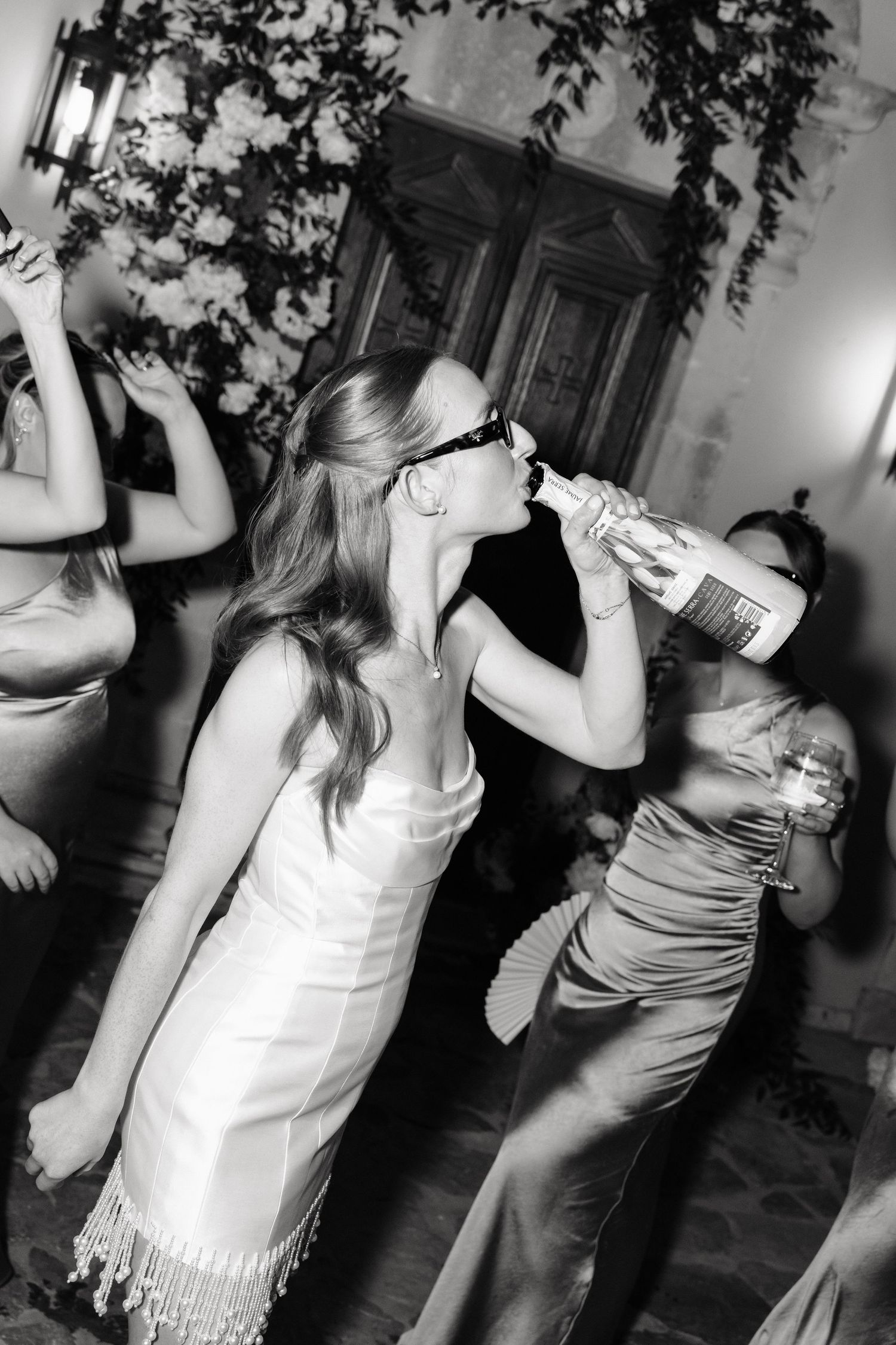 Black and white photo of a bride drinking from a bottle at a wedding reception while guests dance around her.