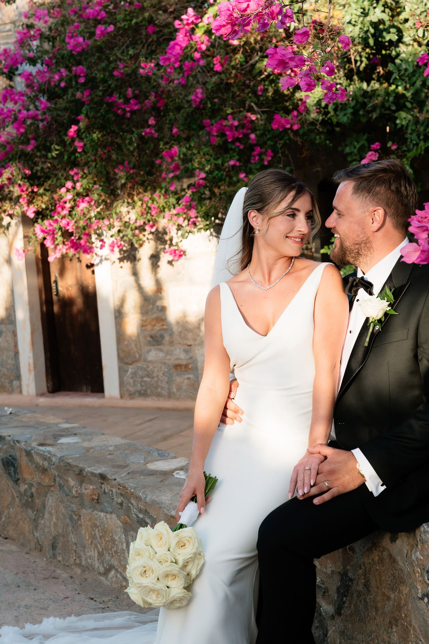 A romantic wedding couple embraces beneath vibrant pink bougainvillea flowers along a stone wall in Mediterranean setting.