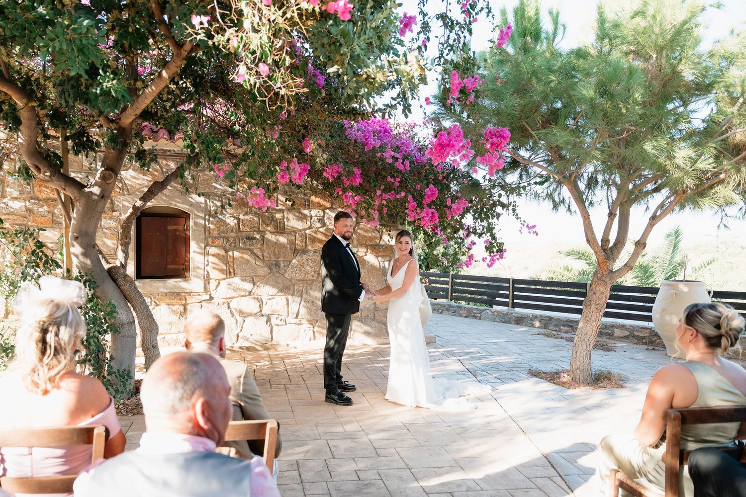 A panoramic view of a Mediterranean terrace with stone walls, potted plants, and pink bougainvillea flowers overhead.