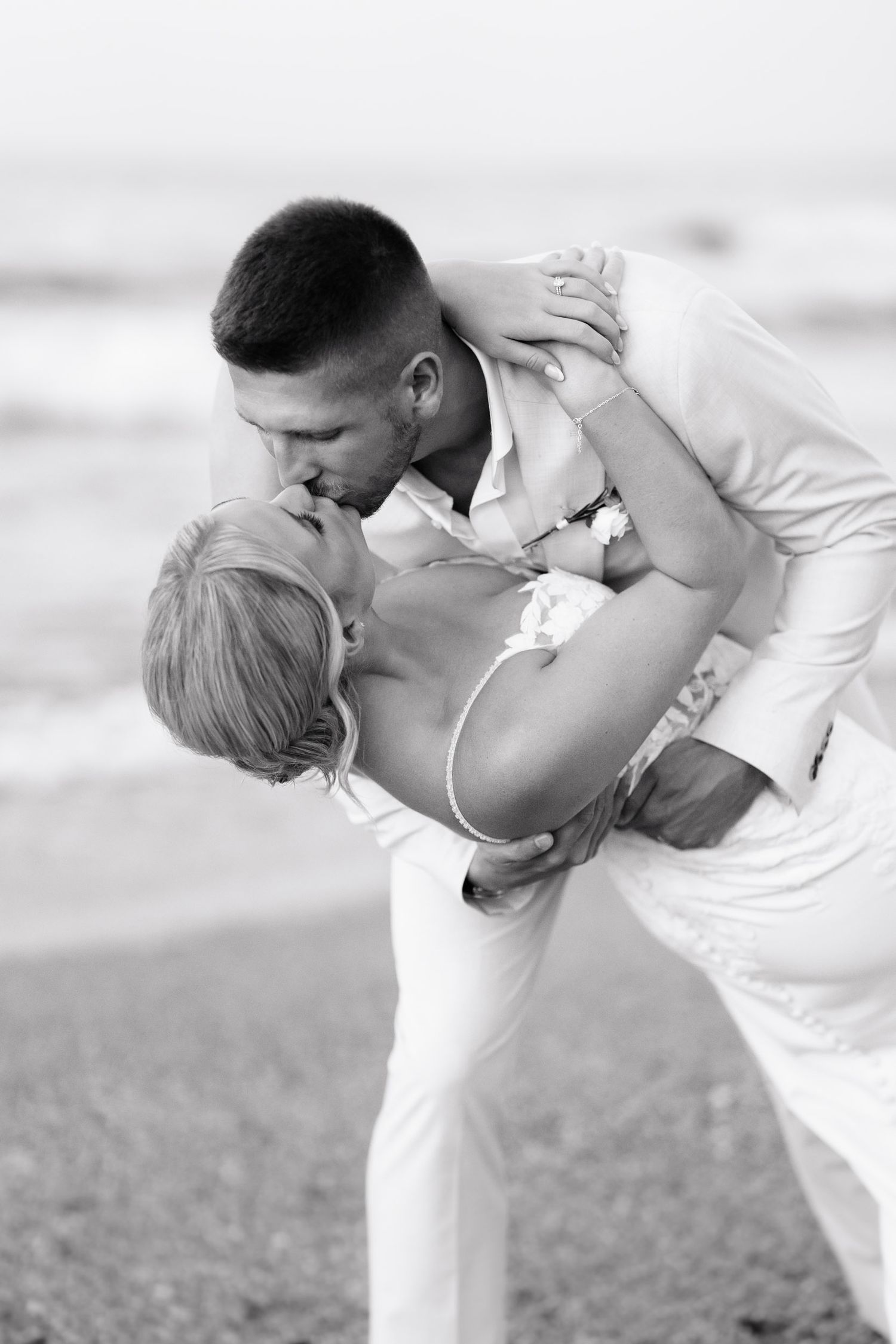 A romantic black and white beach photo of a couple in white clothing sharing a passionate kiss in a dip pose.