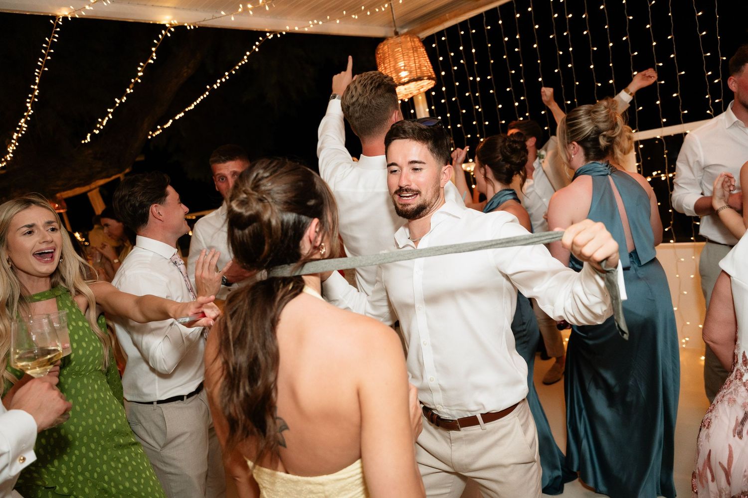 Wedding guests dance and celebrate together under twinkling lights at an outdoor evening reception.