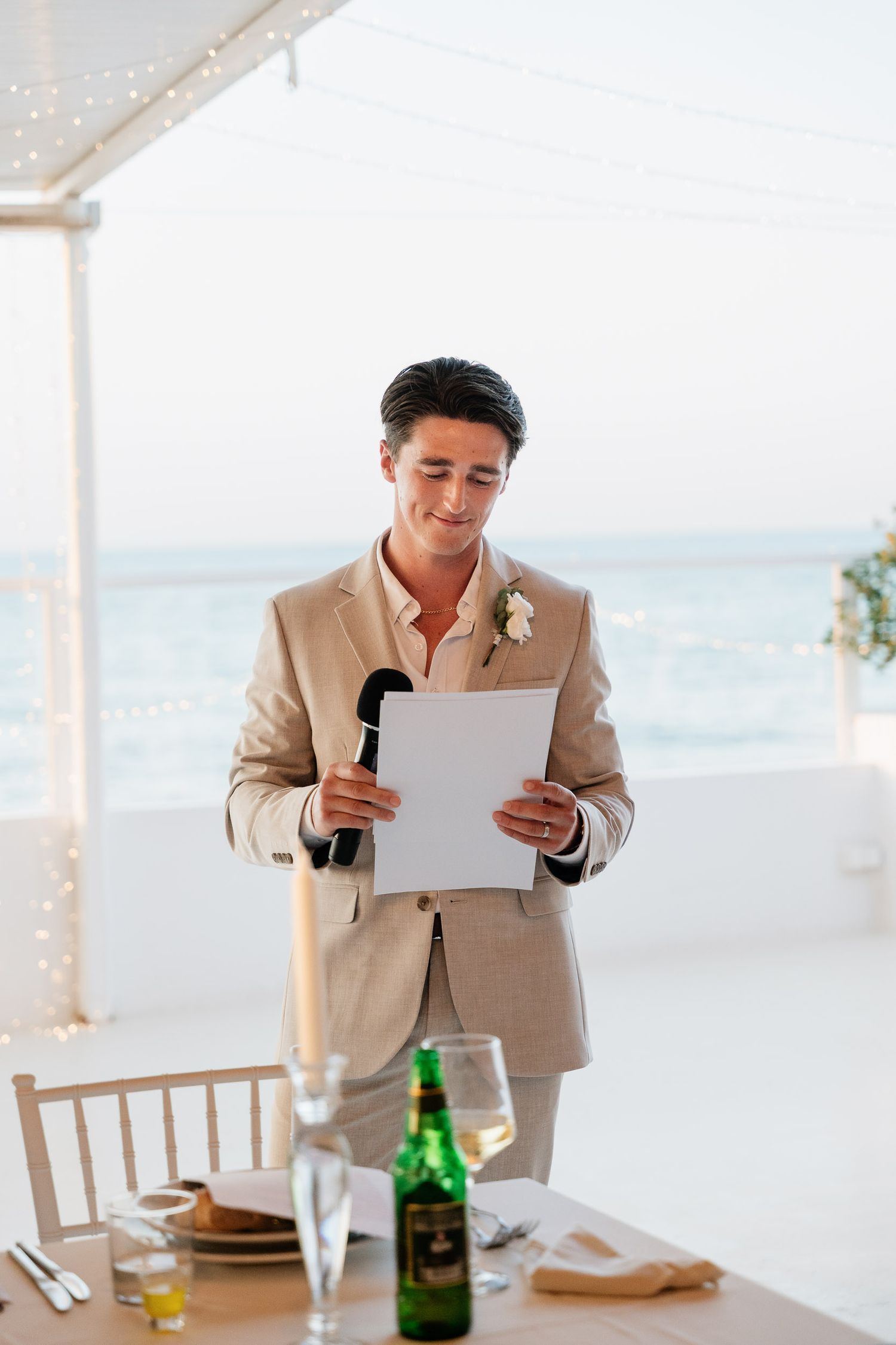 Someone in a beige suit gives a wedding speech at an oceanfront reception venue with drinks on the table.