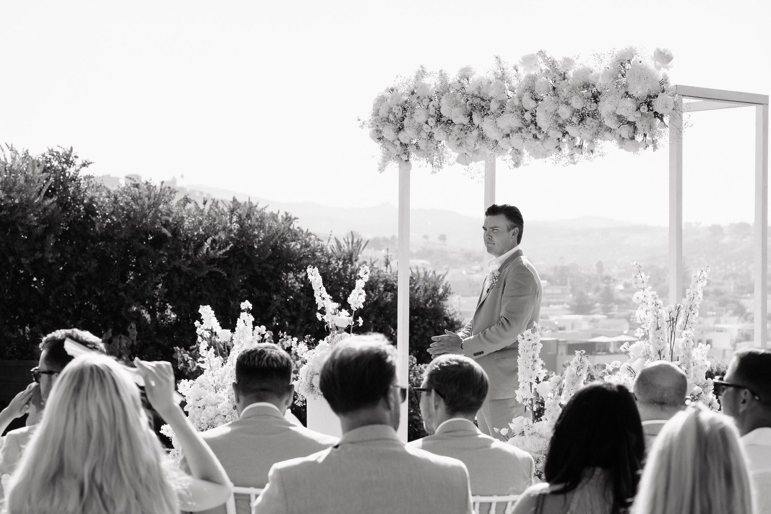 Wedding guests seated outdoors at a ceremony venue with white chairs and decorative floral arrangements.