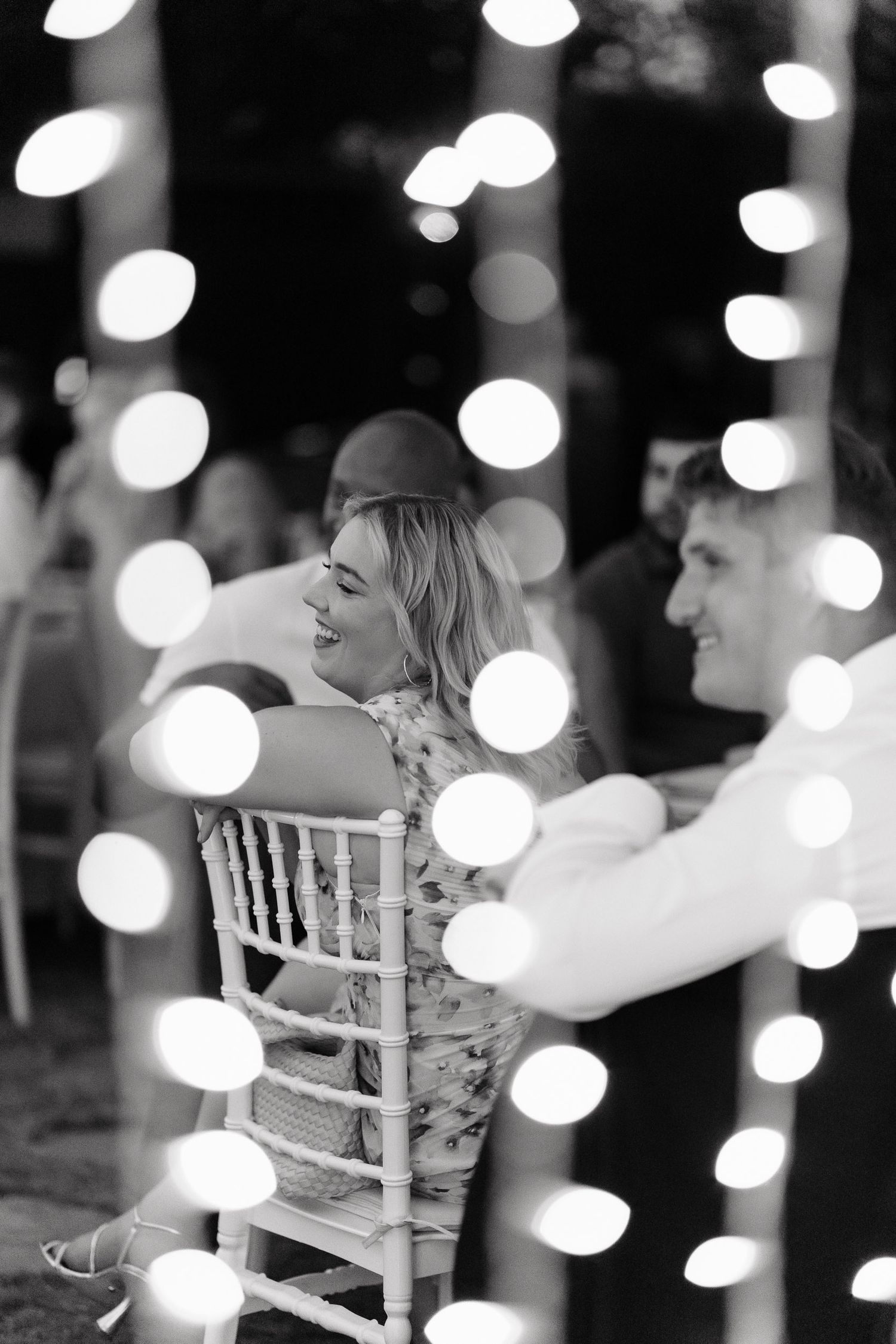 Black and white photo of guests at an event sitting on white chairs with dreamy bokeh lights creating a romantic atmosphere.
