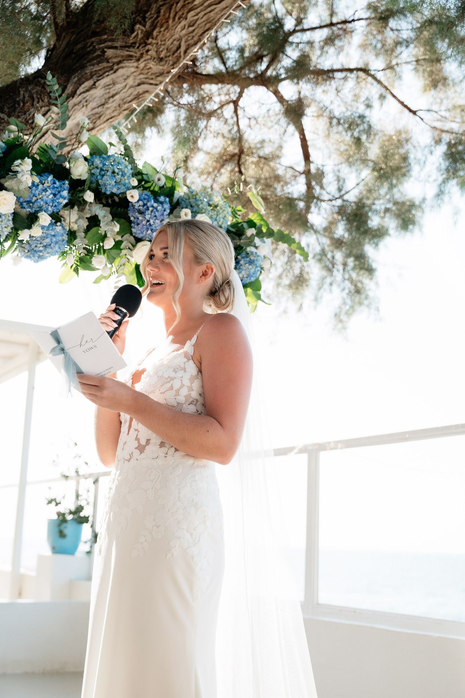 A bride in a white lace dress gives a speech under a floral arch with blue hydrangeas at an outdoor wedding venue.