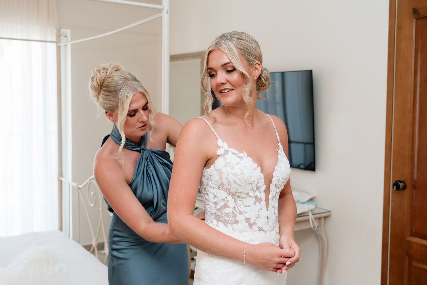 A bride and bridesmaid share an emotional embrace while getting ready in a sunlit room on wedding day.