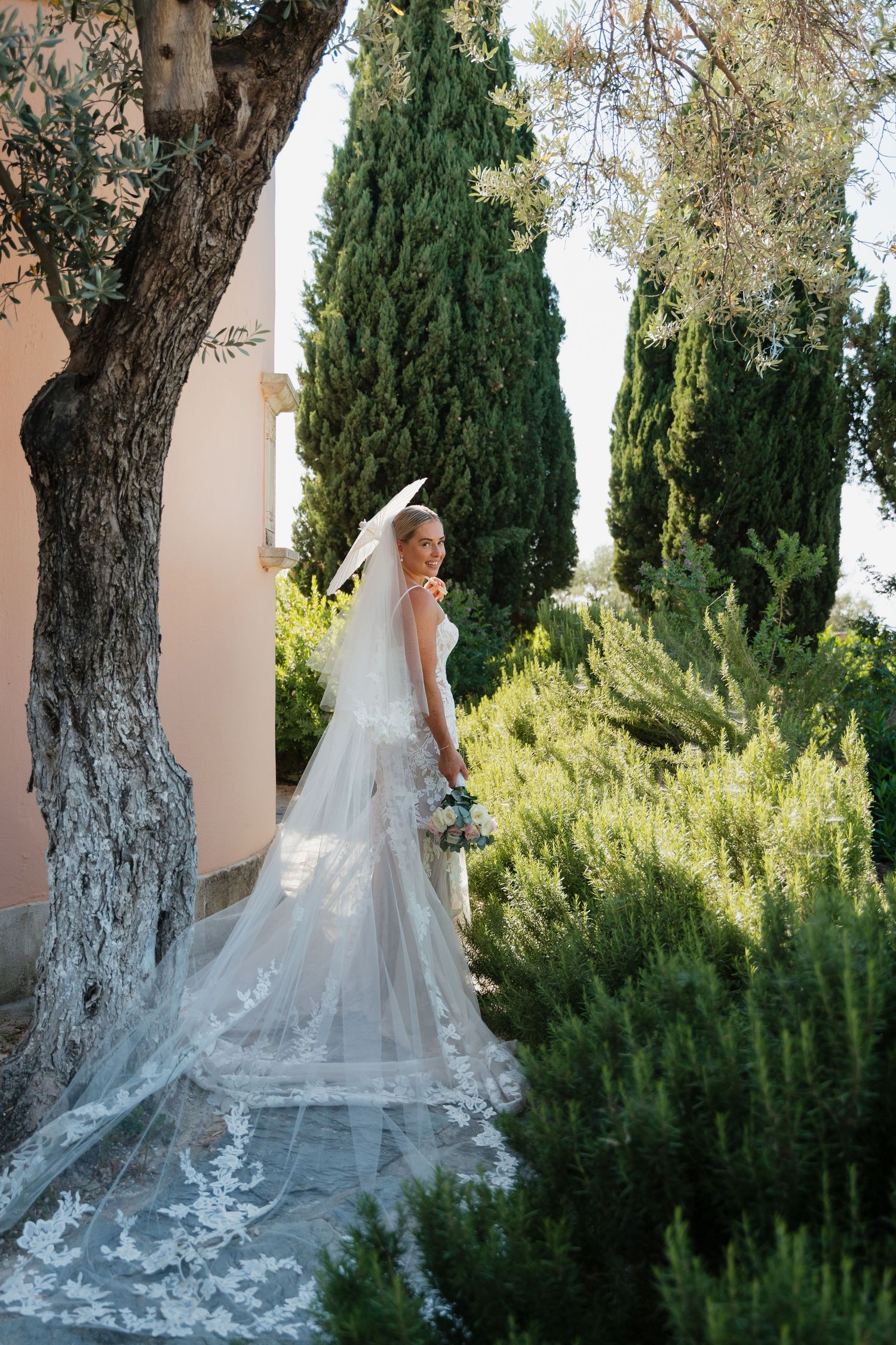 A flowing white wedding dress with intricate lace details cascades elegantly against lush garden greenery and cypress trees.