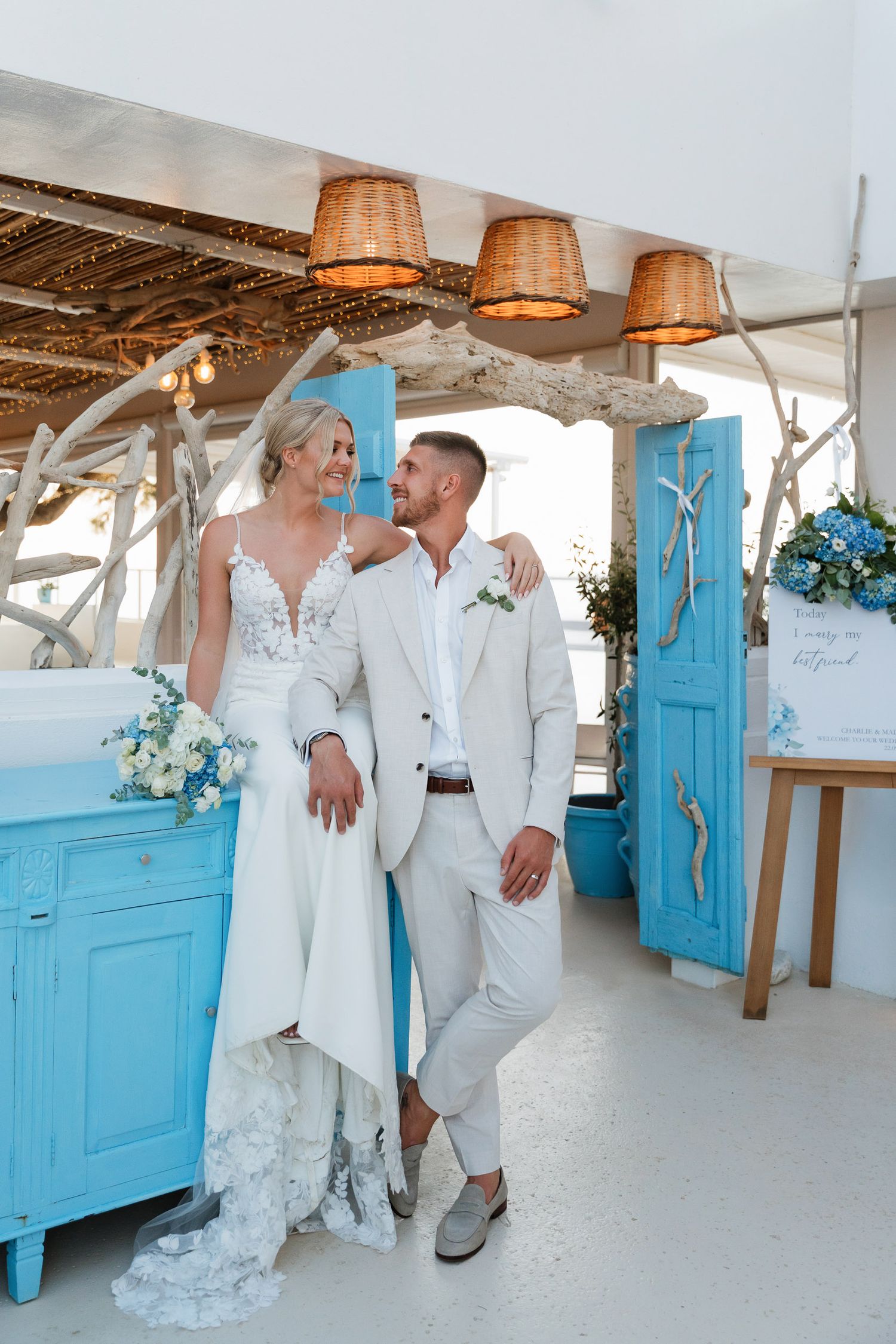 Newlywed couple poses in white attire against rustic beach decor with turquoise blue doors and barrel lamp shades.