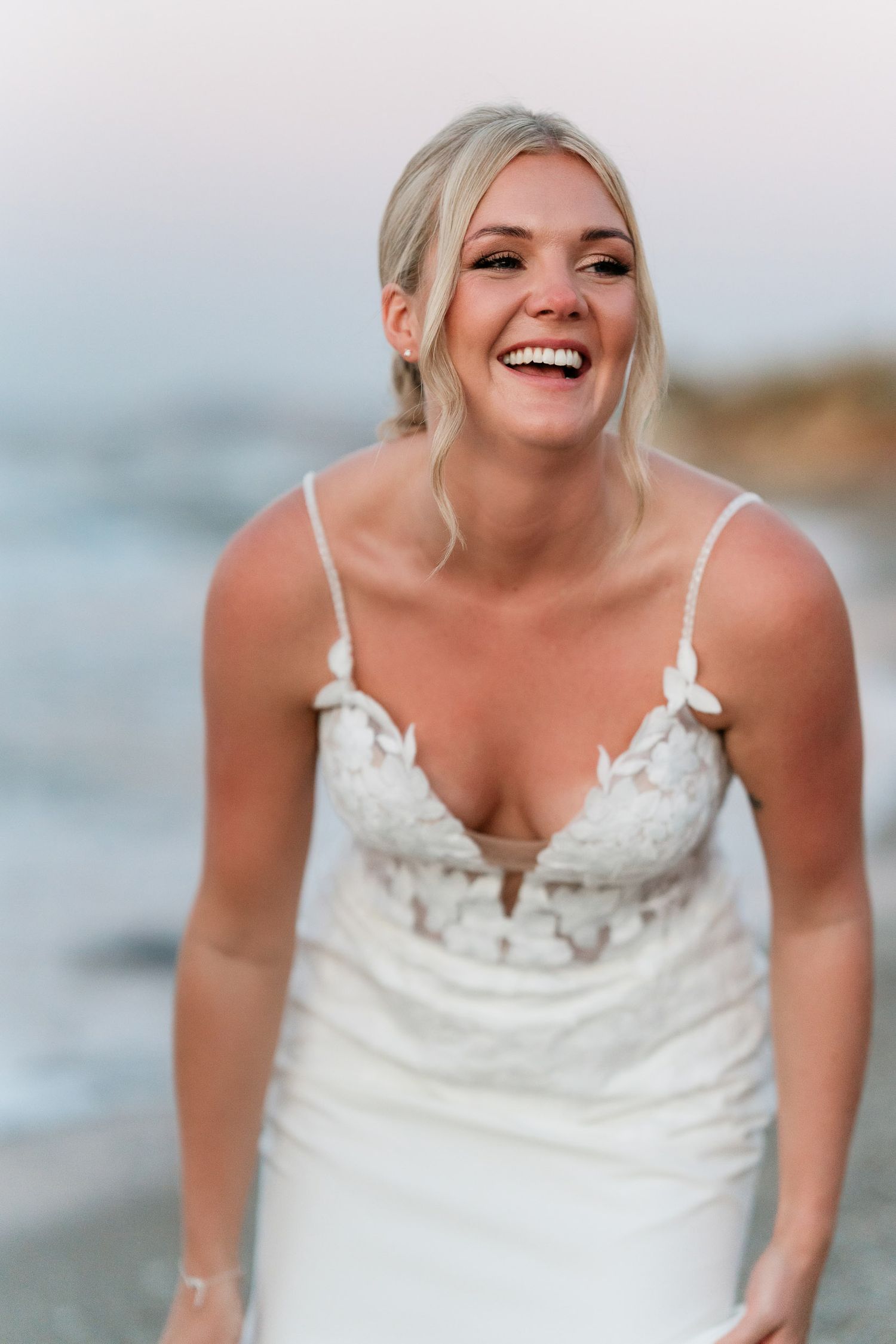 A bride in a white wedding dress laughs joyfully during a beach photoshoot with waves in the background.
