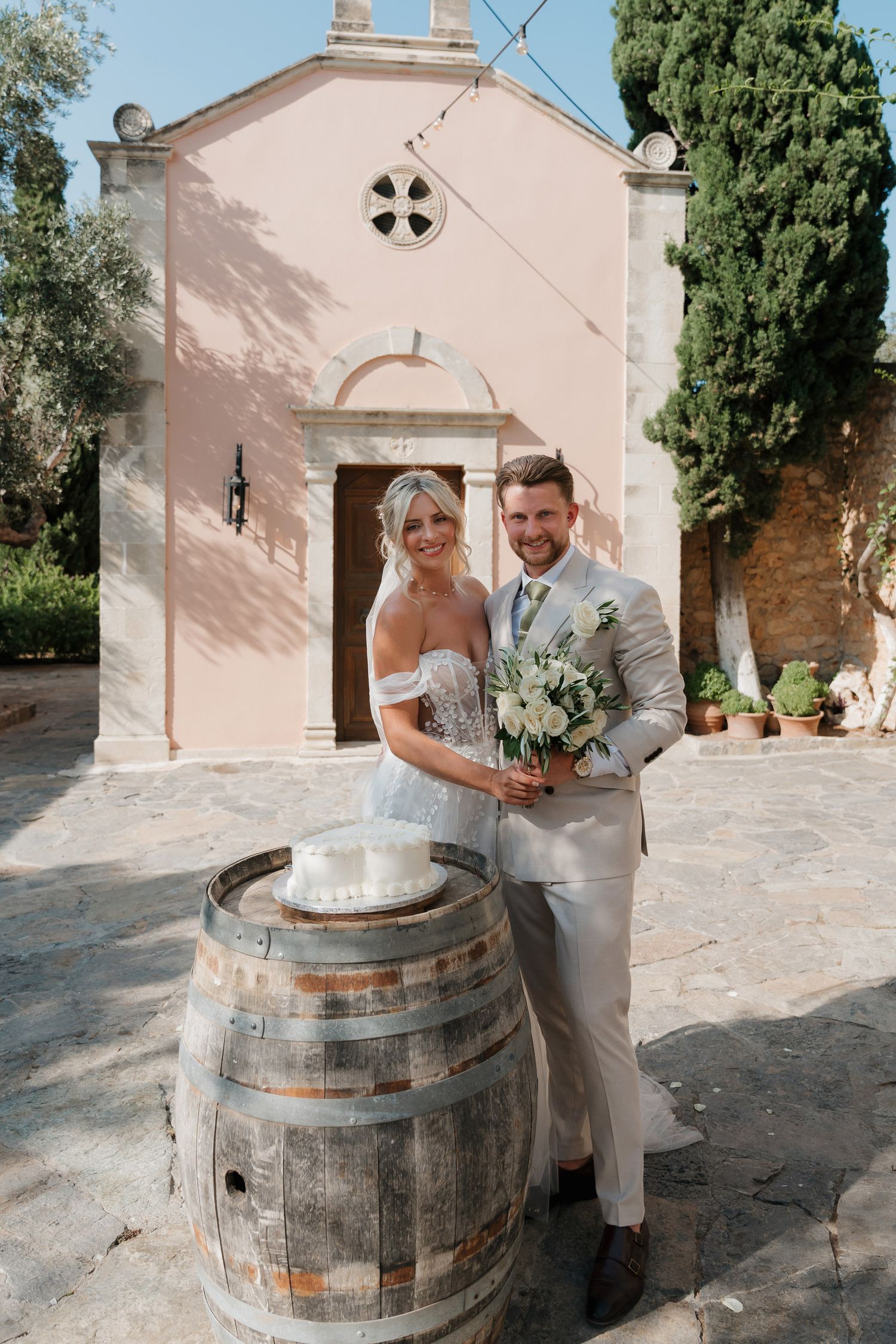 Couple in light-colored attire posing with a rustic wine barrel outside a Mediterranean church with pink walls and cypress trees.