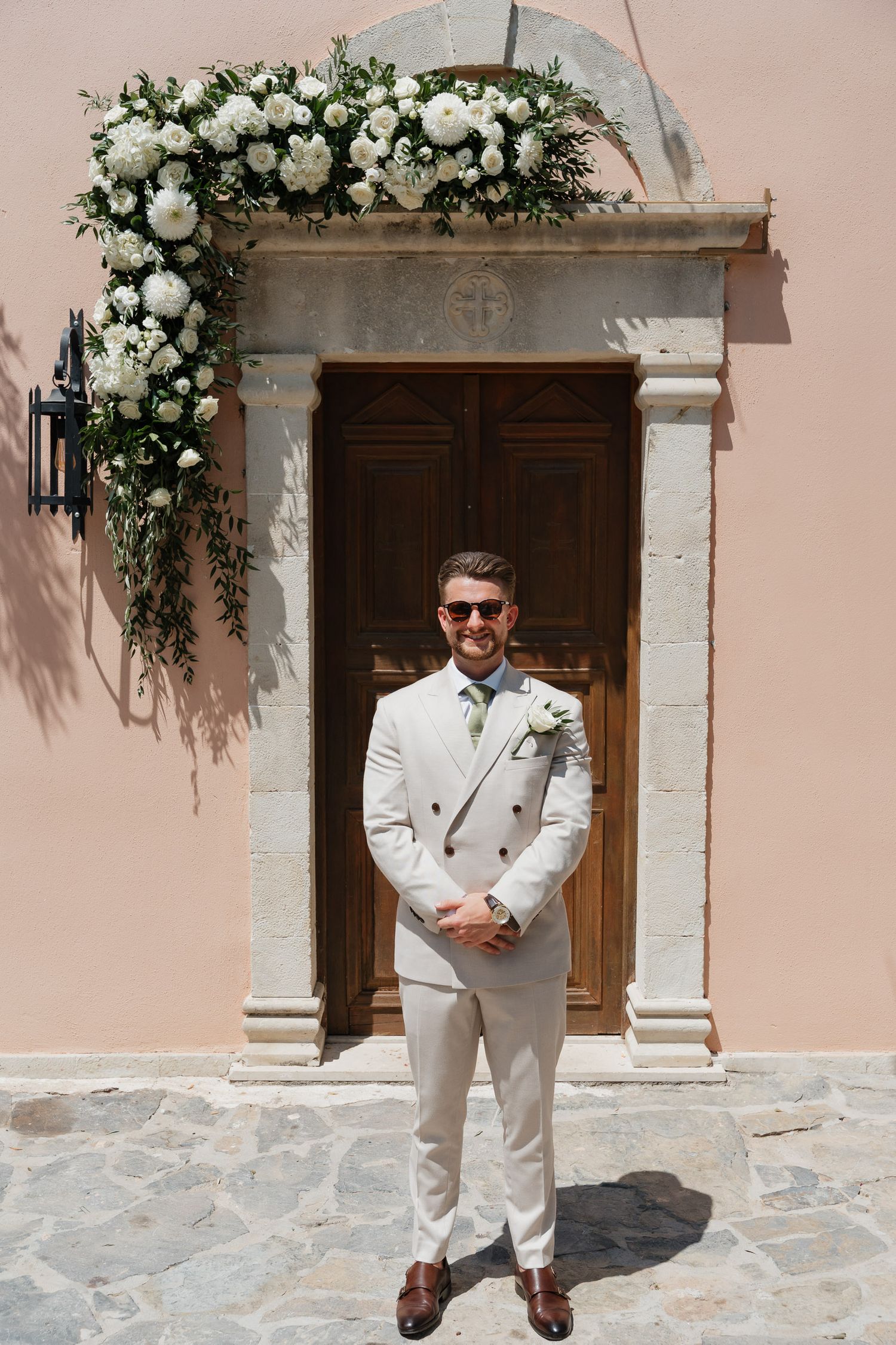 Elegant floral arch with white roses decorates a wooden doorway entrance with stone columns and peach-colored walls.