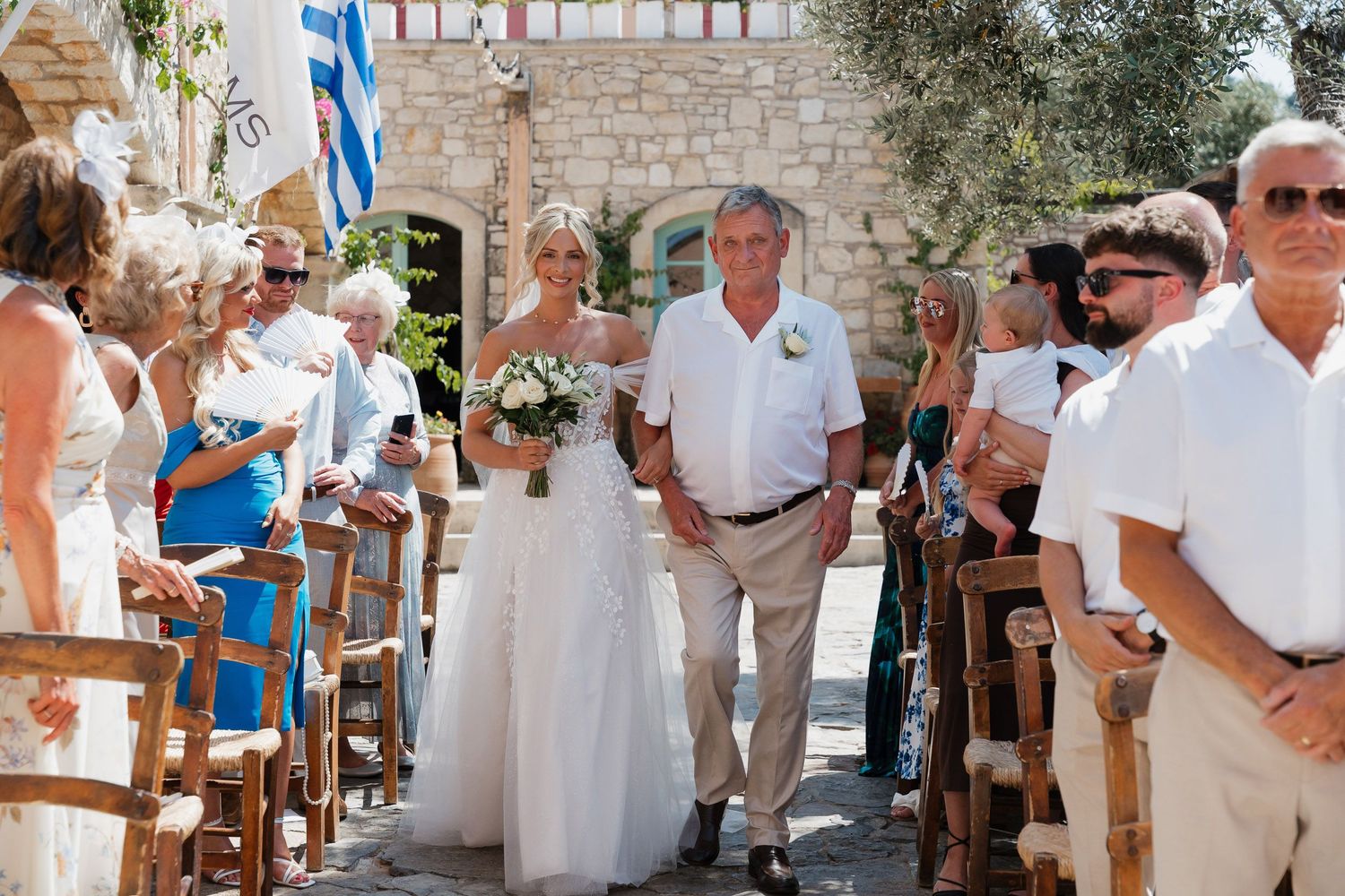 People in white clothing gather for a celebration at an outdoor venue with stone walls and olive trees.