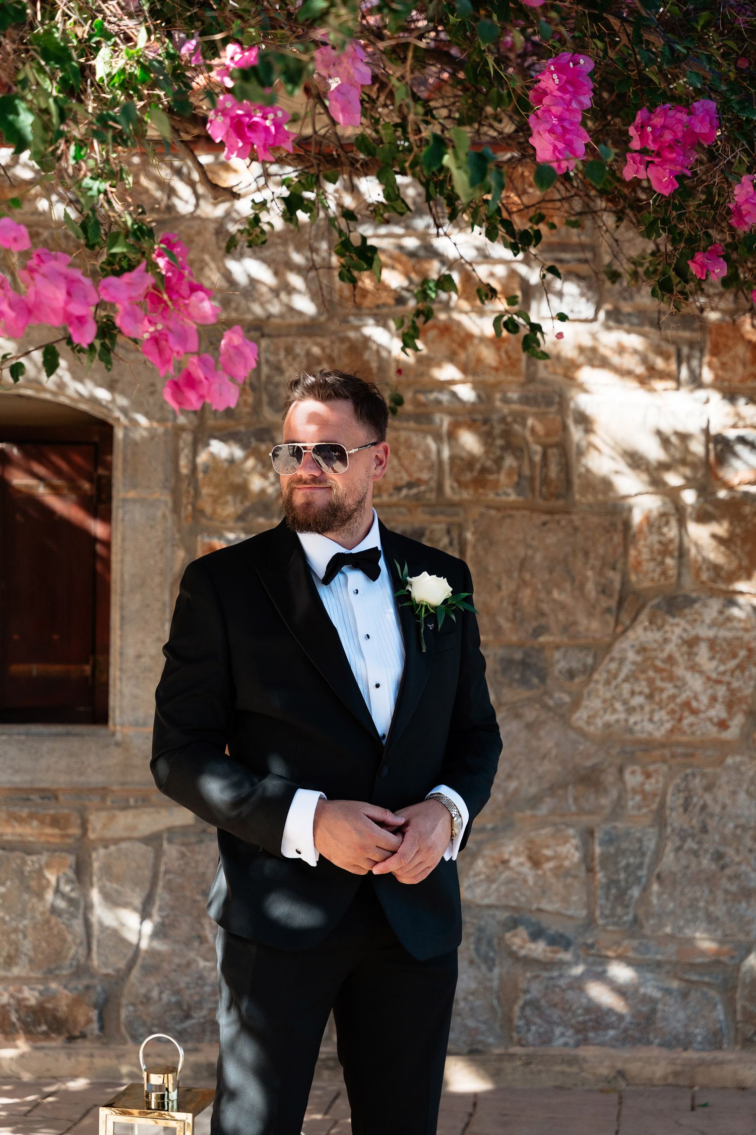A person in a black tuxedo and bow tie stands against a stone wall with vibrant pink bougainvillea flowers above.
