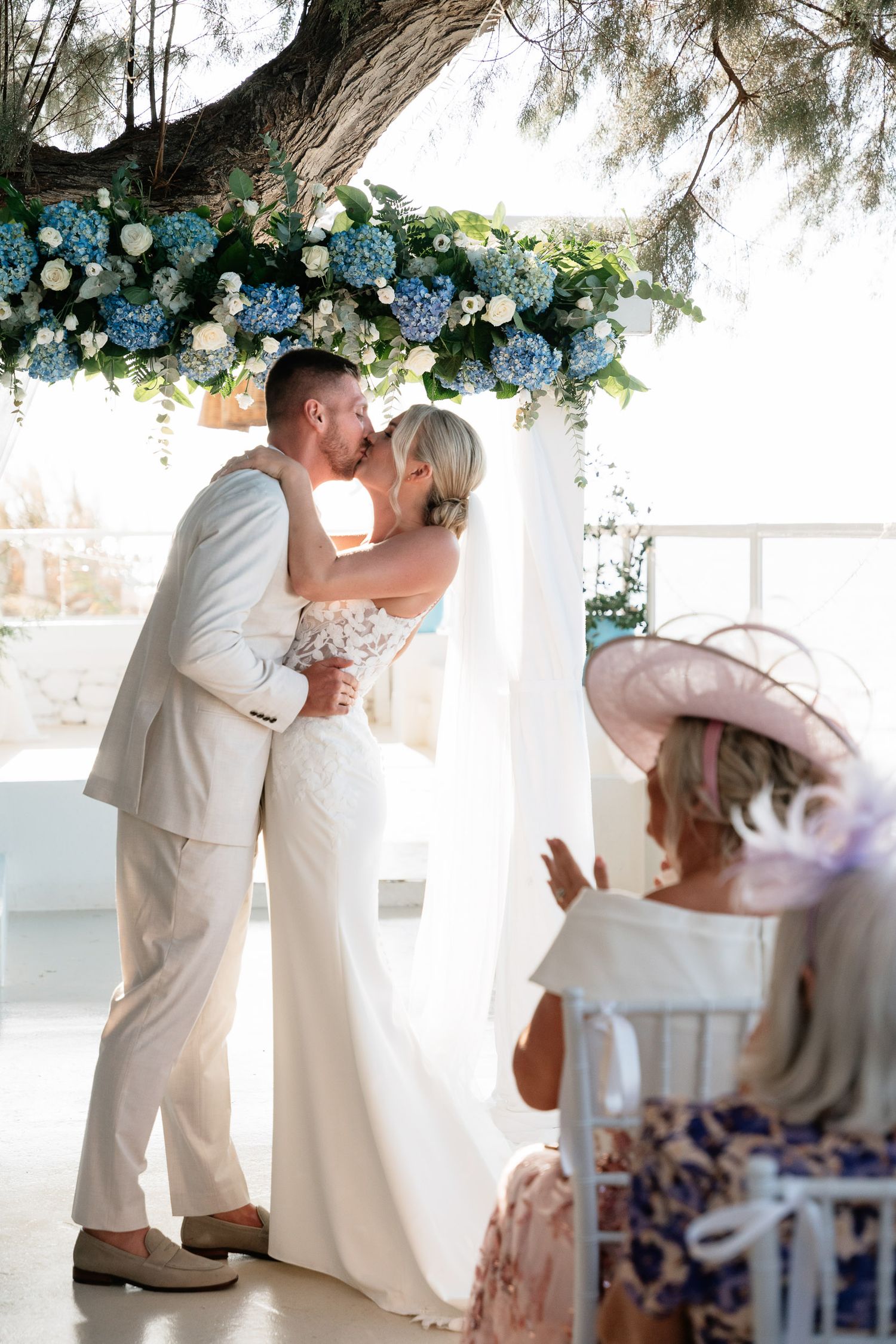 A romantic wedding couple shares intimate moments under floral decorations in a bright white venue.
