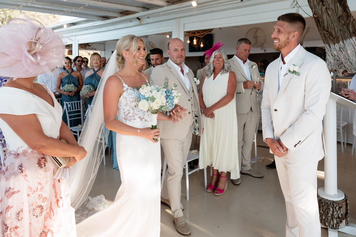 A couple exchanges vows during a beachside wedding ceremony with guests looking on in white and floral attire.