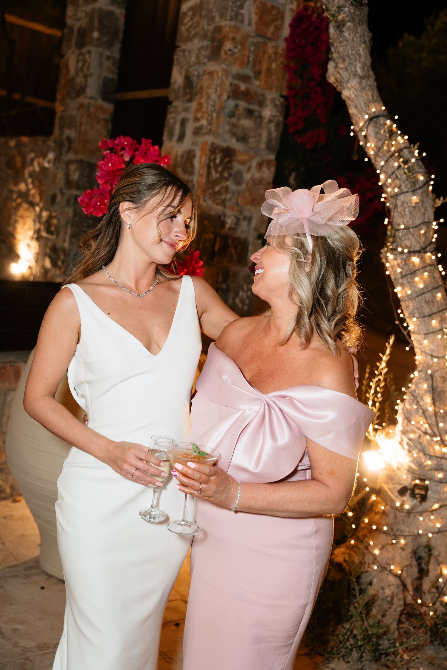 Two people in white and pink dresses share a joyful moment dancing together at an elegant stone-walled venue with twinkle lights.