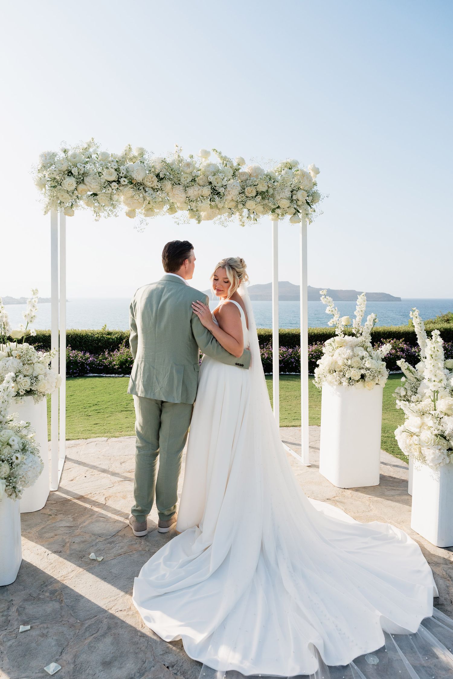 A couple shares a moment under a white floral arch at their outdoor wedding ceremony with mountain views in the background.