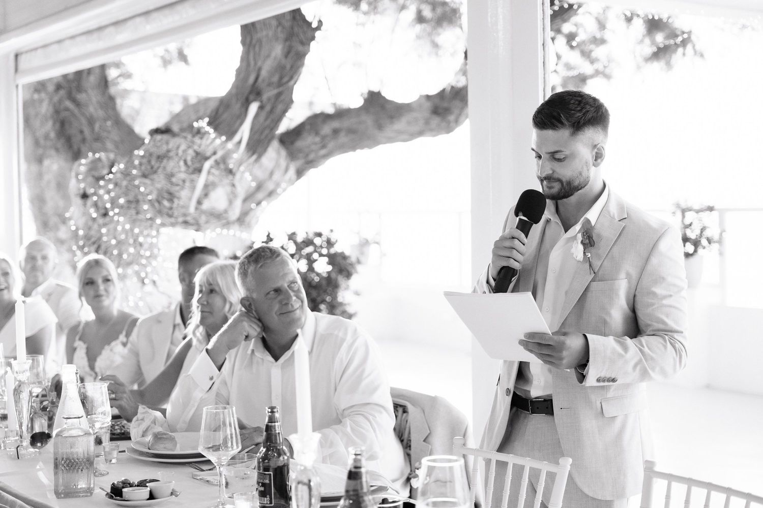 A black and white photograph of a wedding reception speech being delivered under an outdoor tent or pavilion.