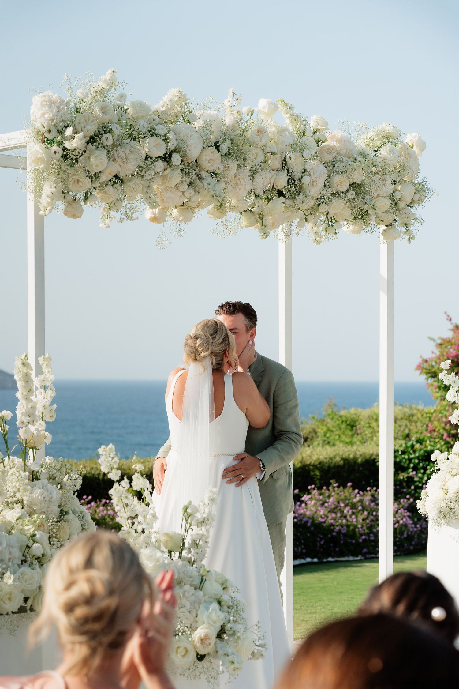 A romantic outdoor wedding ceremony overlooking the ocean with a white floral arch and guests in attendance.