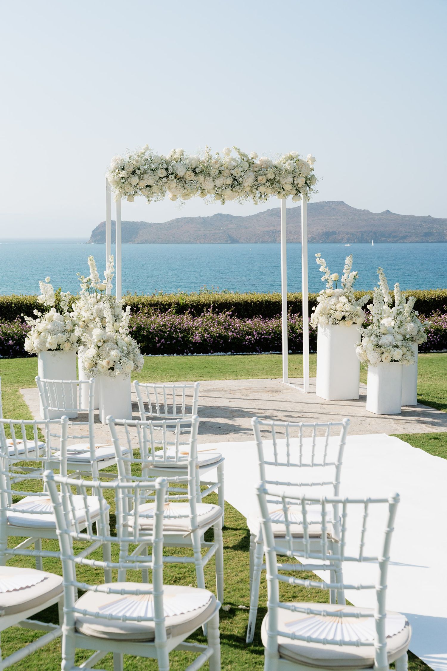 White chairs arranged for an outdoor wedding ceremony with ocean and mountain views create an elegant seaside setting.