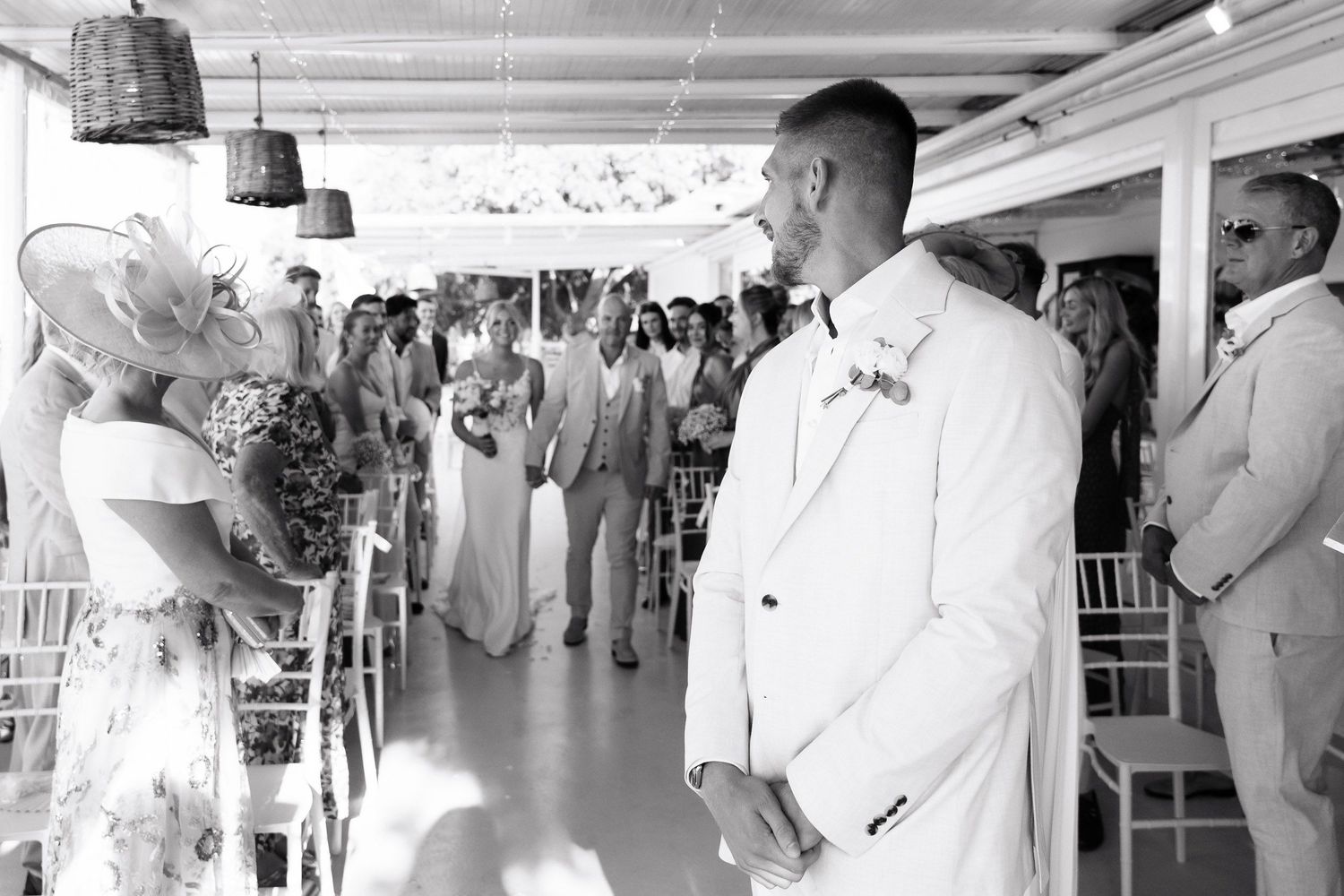 A black and white photograph captures a wedding ceremony moment with guests seated in rows at an elegant venue.