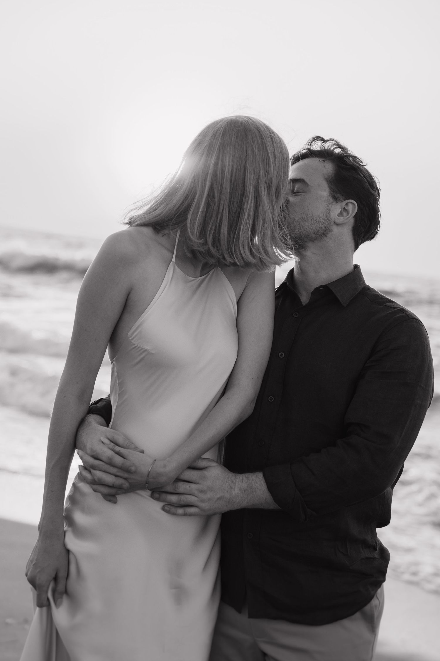 A romantic black and white photo series shows a couple embracing and kissing on a beach at sunset.