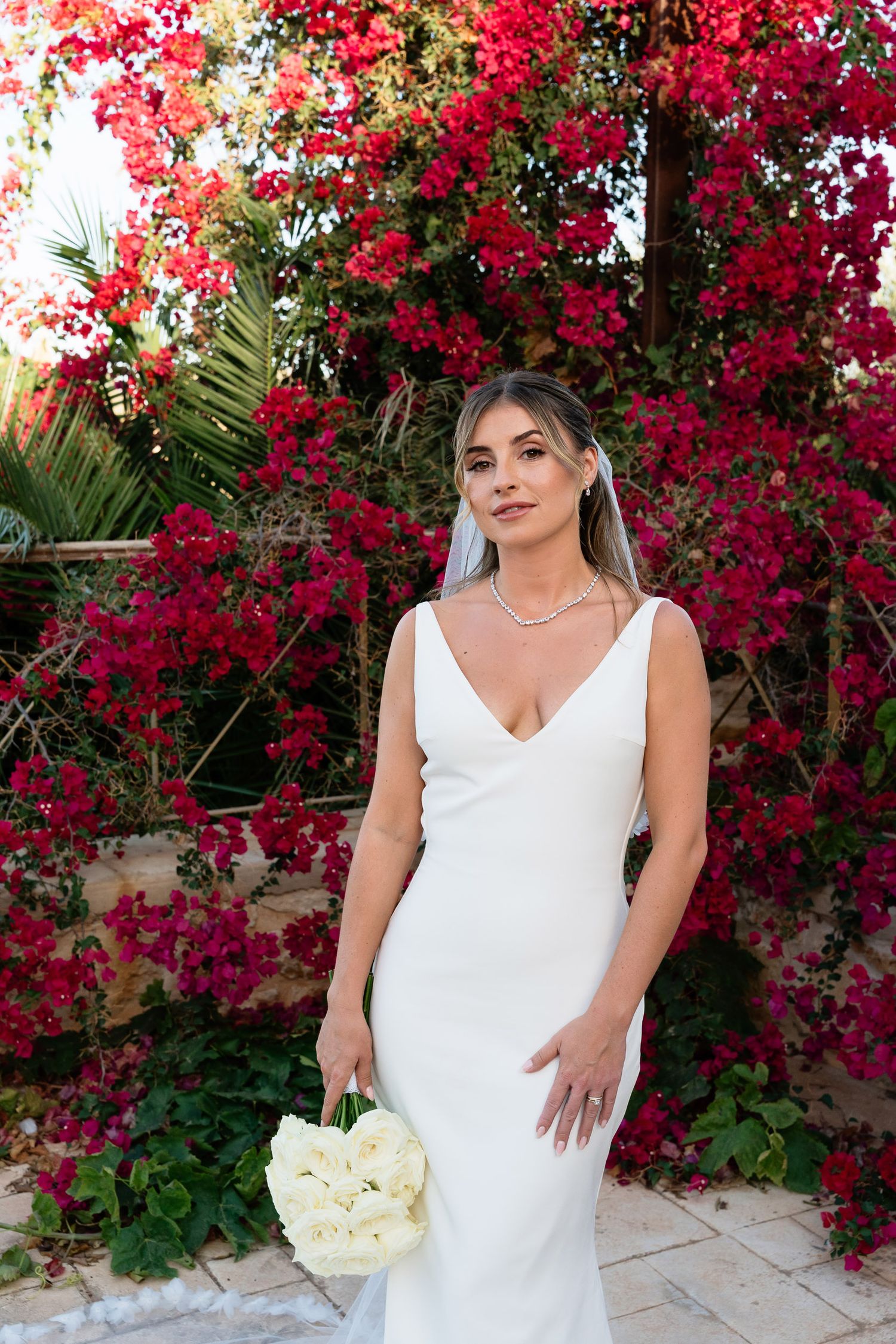 A bride in a sleek white wedding dress holds a white bouquet against a backdrop of vibrant red bougainvillea flowers.