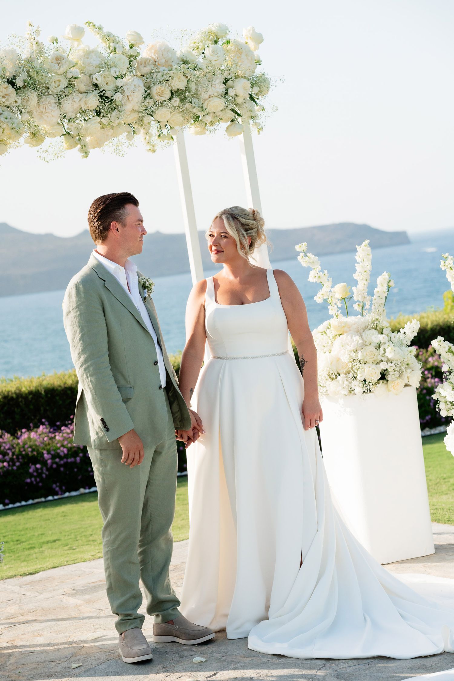 A couple shares a joyful moment under a white floral arch during their seaside wedding ceremony in Greece.