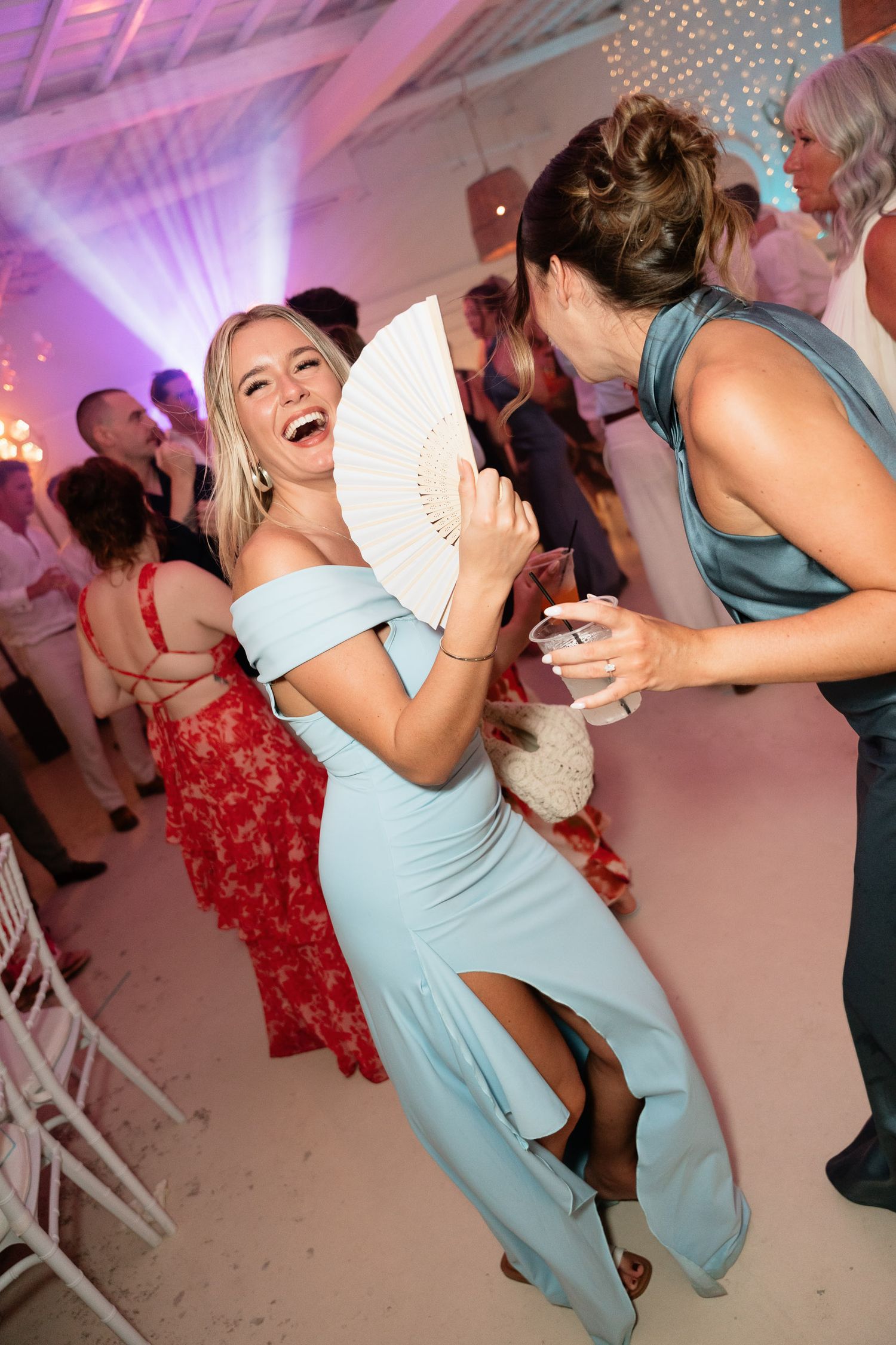Wedding guests dance and celebrate on a pink-lit dance floor with people wearing light blue and red dresses.