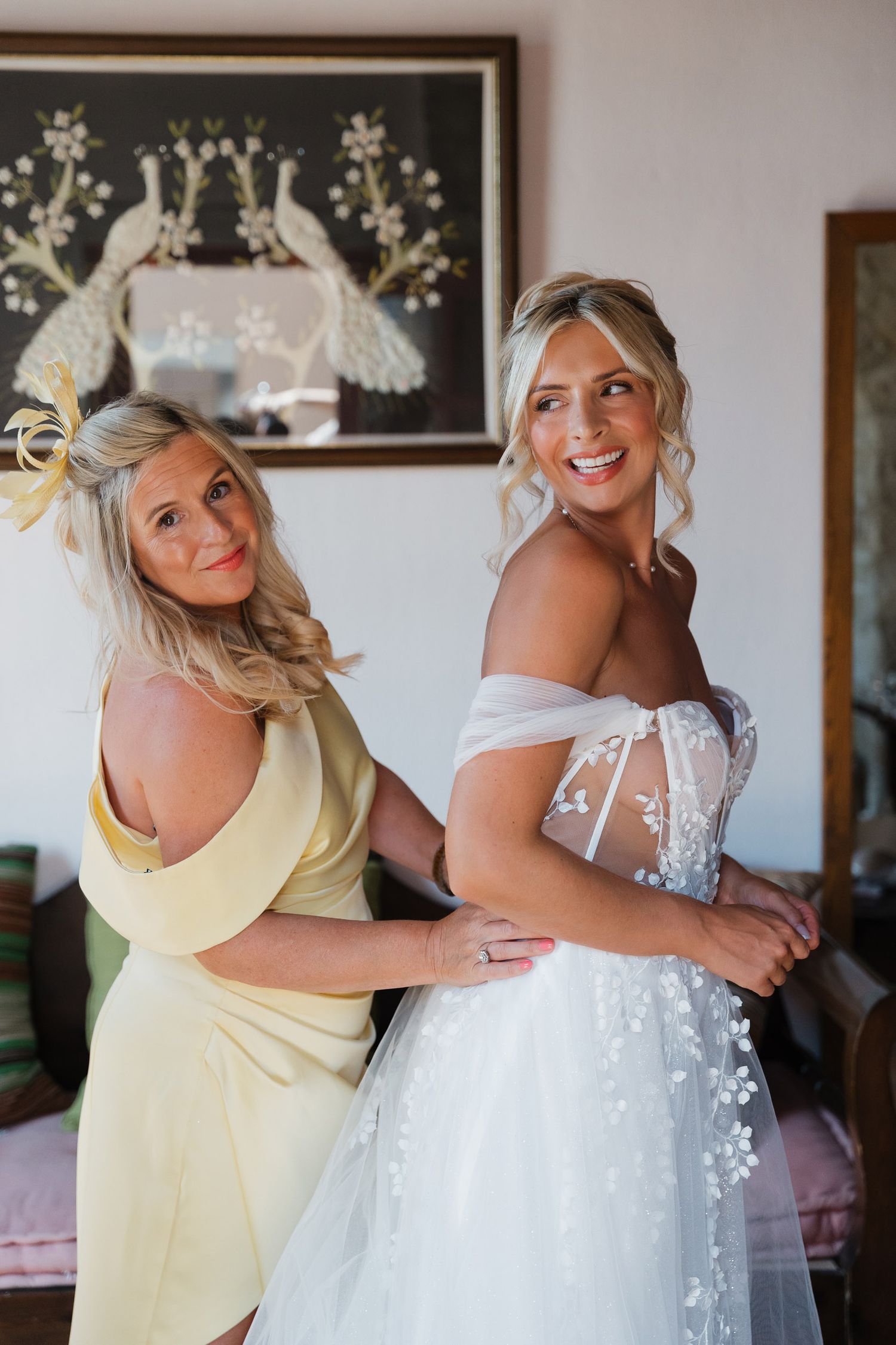 Joyful wedding preparation moment as bridesmaid in yellow dress helps with final touches on bride's elegant gown.