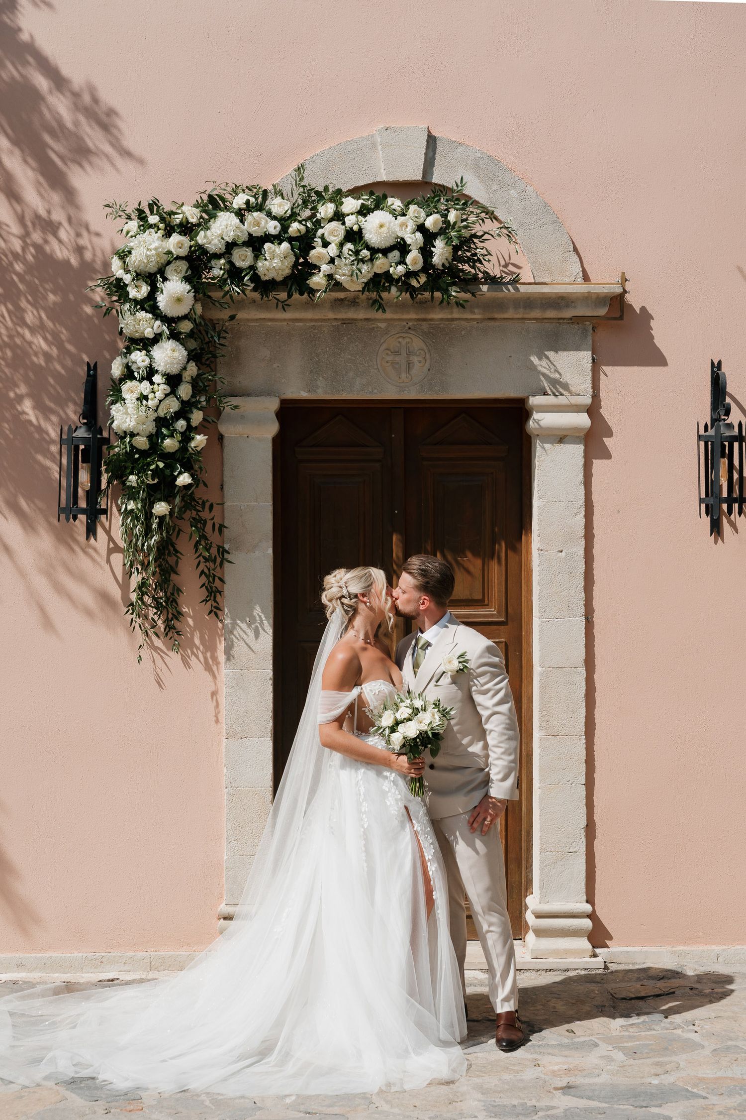 A romantic wedding couple shares an intimate moment in front of a rustic wooden doorway adorned with white floral arrangements.