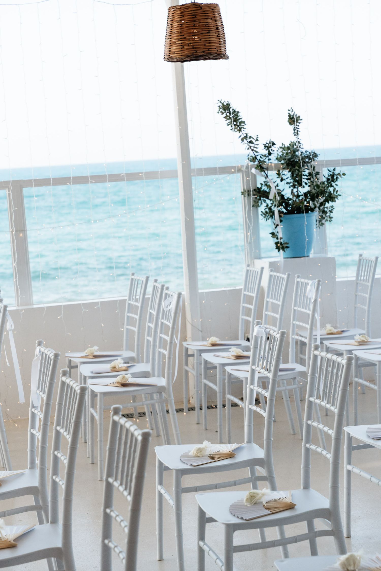 White chairs and tables arranged at an oceanfront restaurant with woven basket pendant lights and turquoise water views.