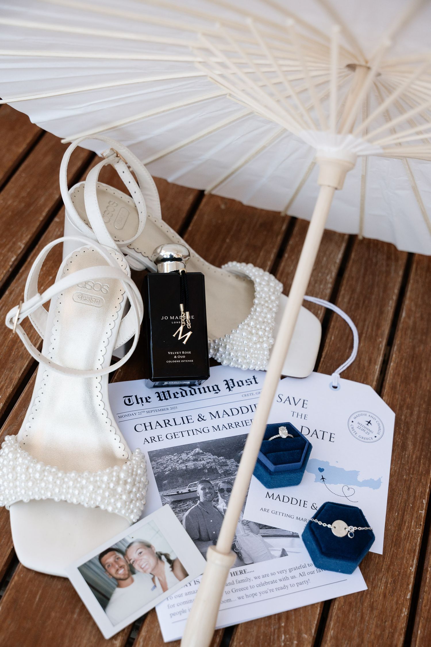 Wedding details including white heels, parasol, perfume bottle and invitation cards arranged on wooden surface.