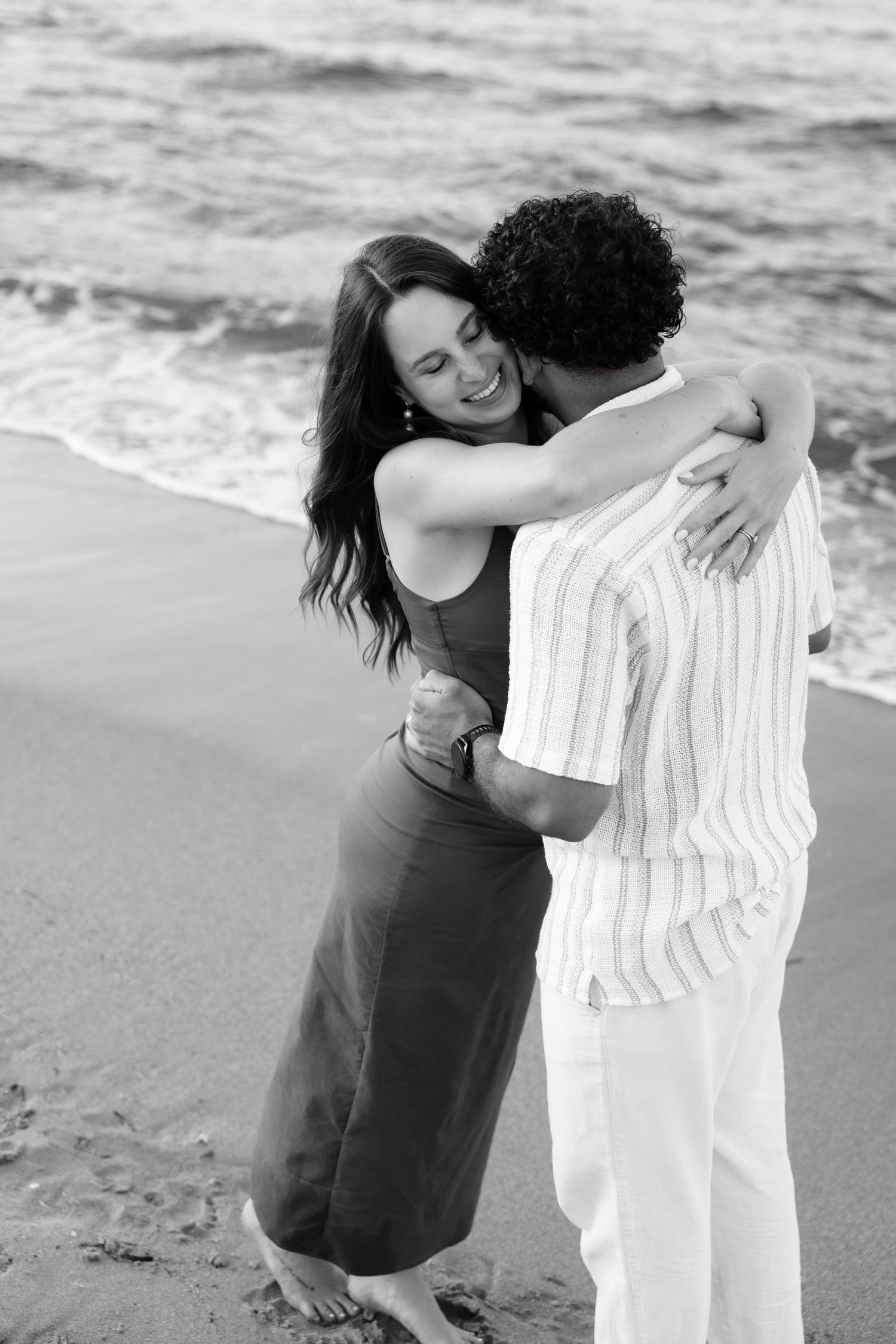 A couple embraces on a beach in black and white, with waves crashing in the background.