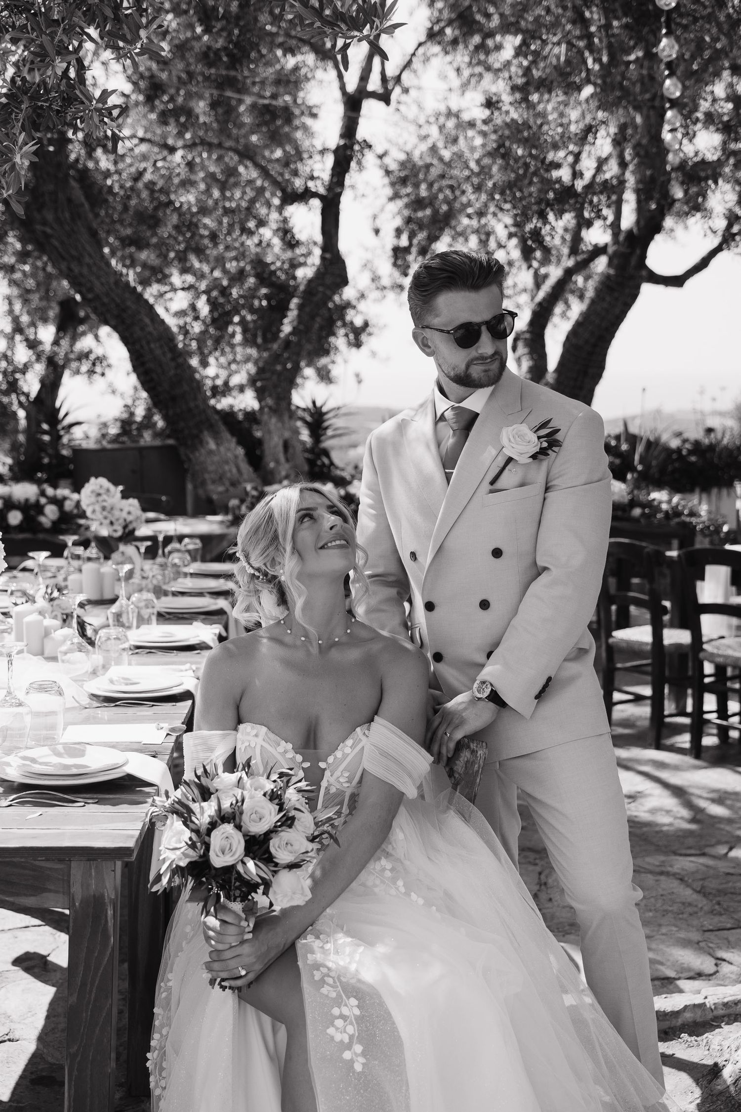 Romantic outdoor wedding scene with couple in white attire dancing and embracing under trees at a garden reception.
