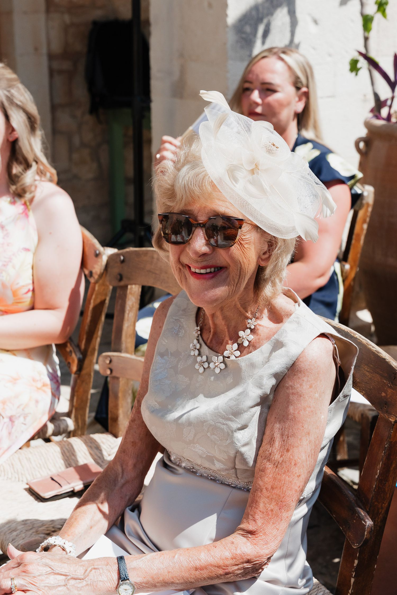 Elderly woman in white dress and sunhat laughs joyfully while sitting in wooden chair at outdoor gathering.