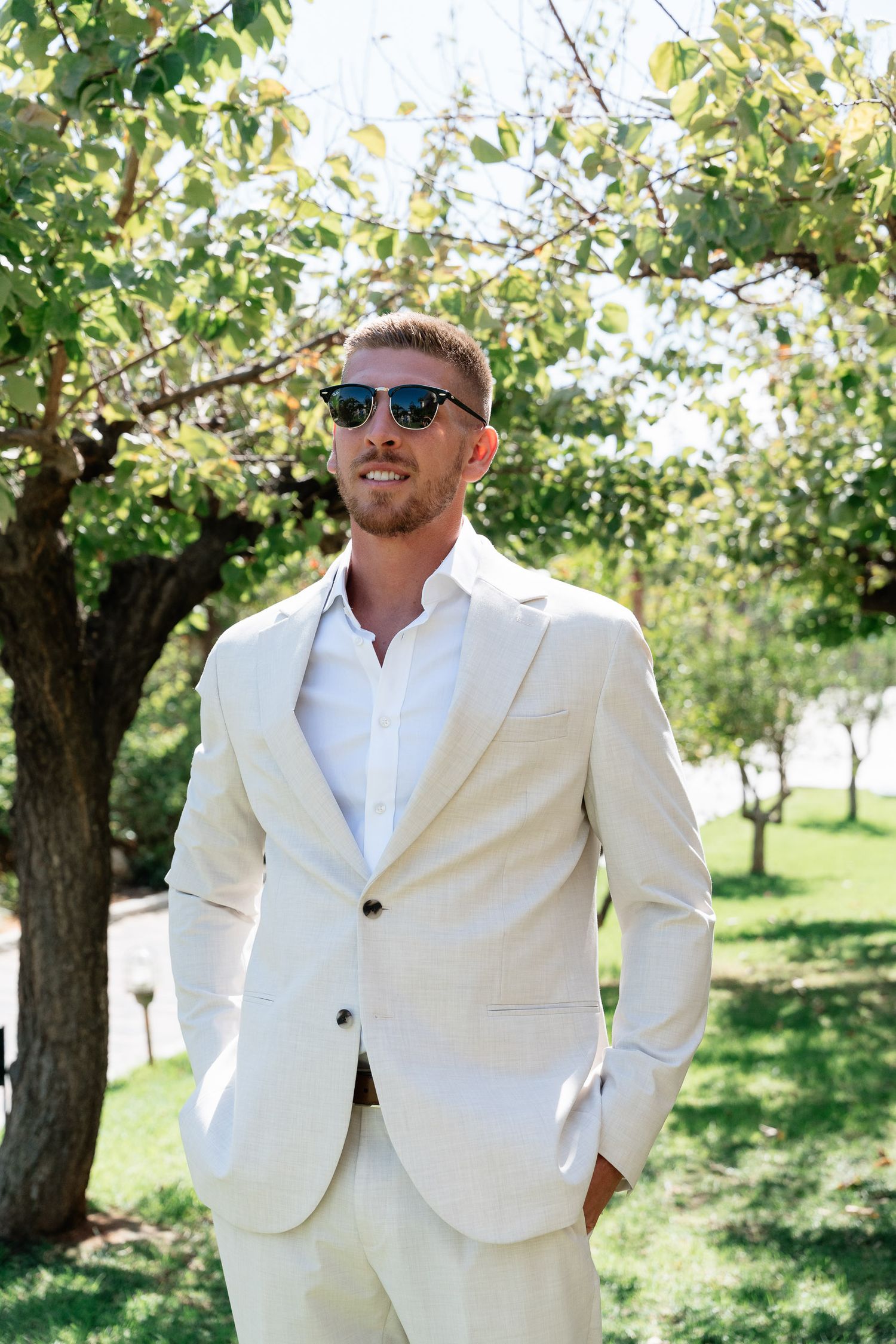 A person in a crisp white summer suit and sunglasses poses outdoors in a garden setting with trees in the background.