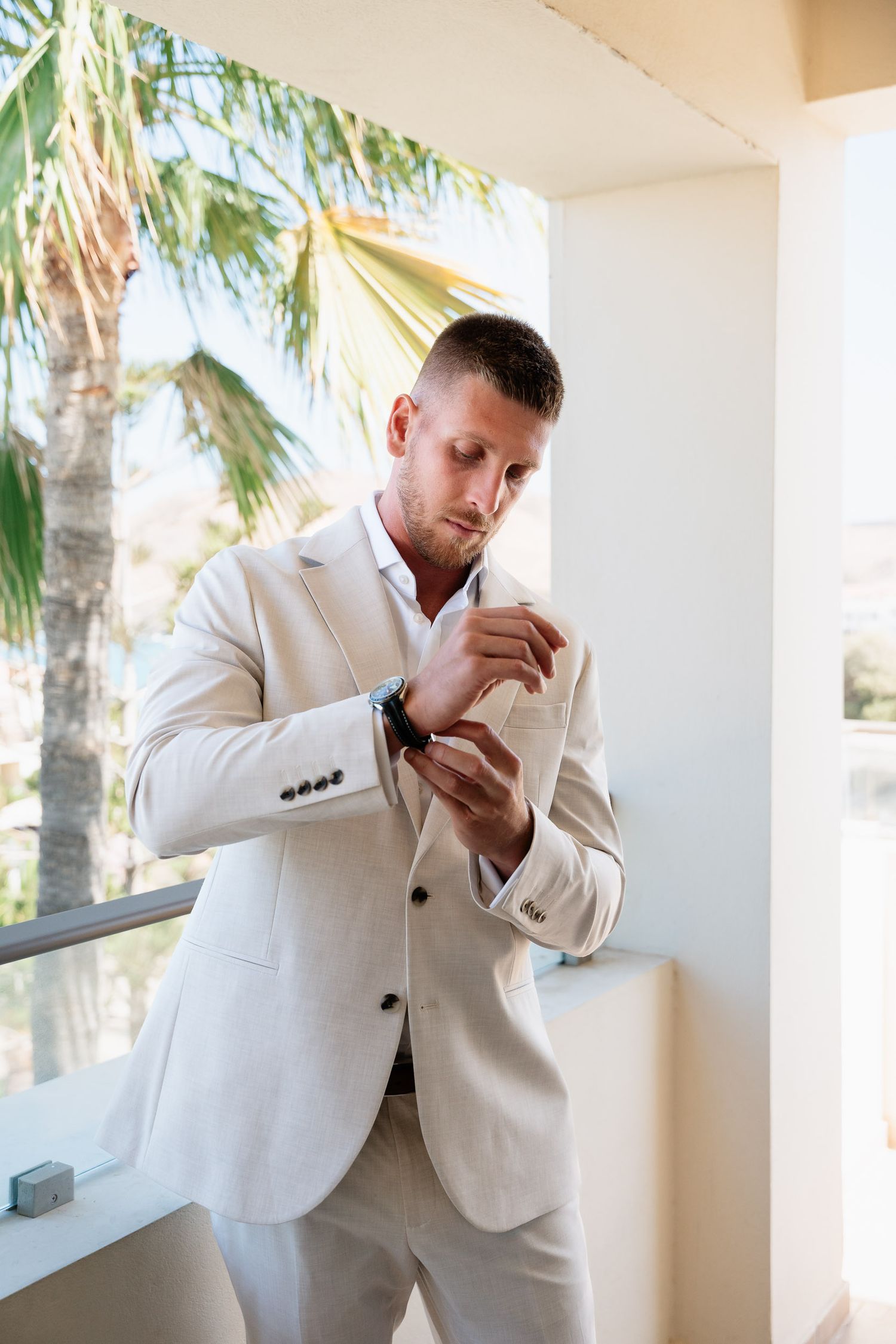 A person in a light colored suit adjusts their cuff while standing on a balcony with palm trees in the background.