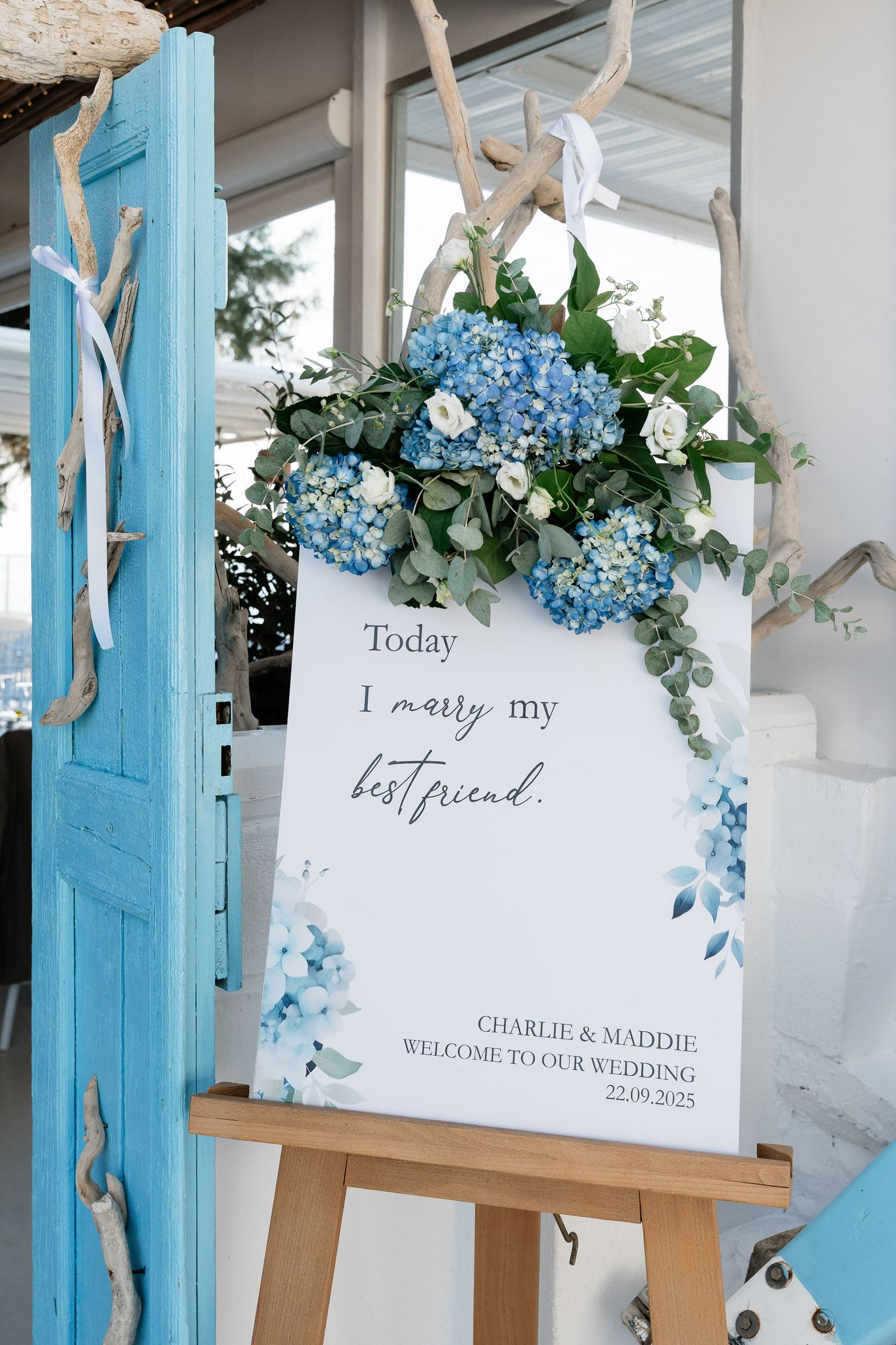 Wedding welcome sign decorated with blue hydrangeas and white flowers against a turquoise door frame.