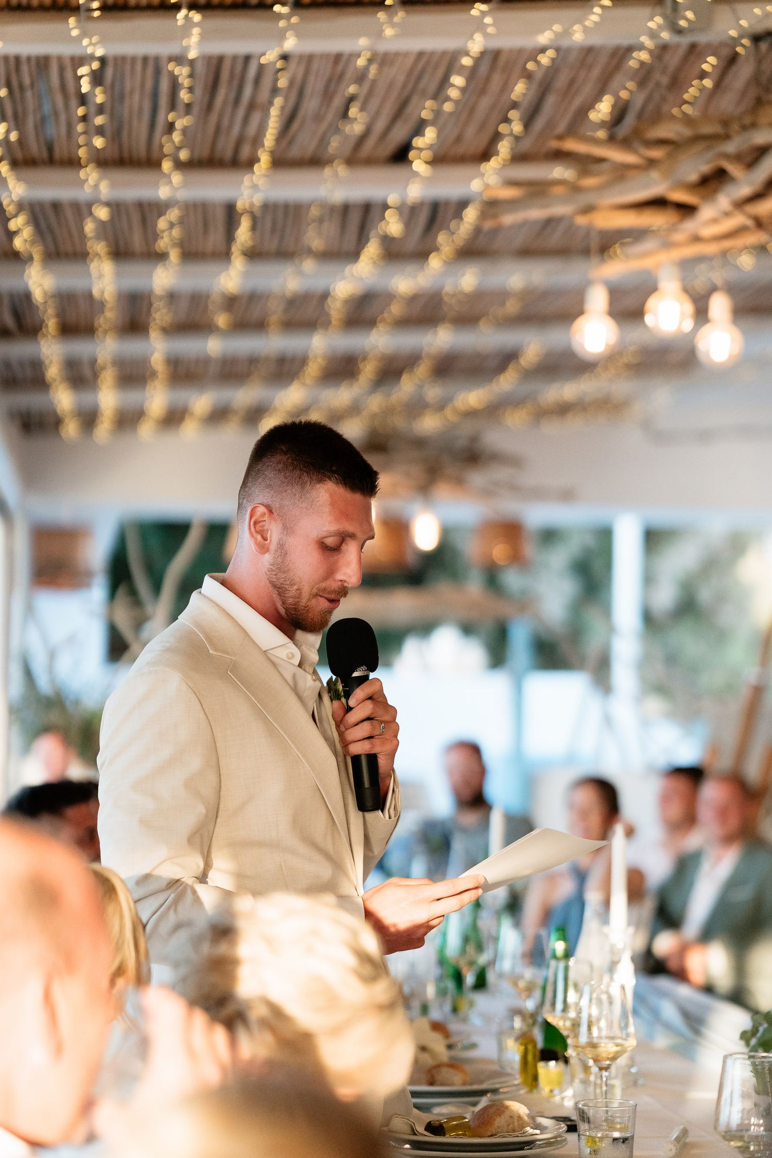 A person in a white dress shirt gives an emotional speech at an elegant indoor reception with string lights overhead.