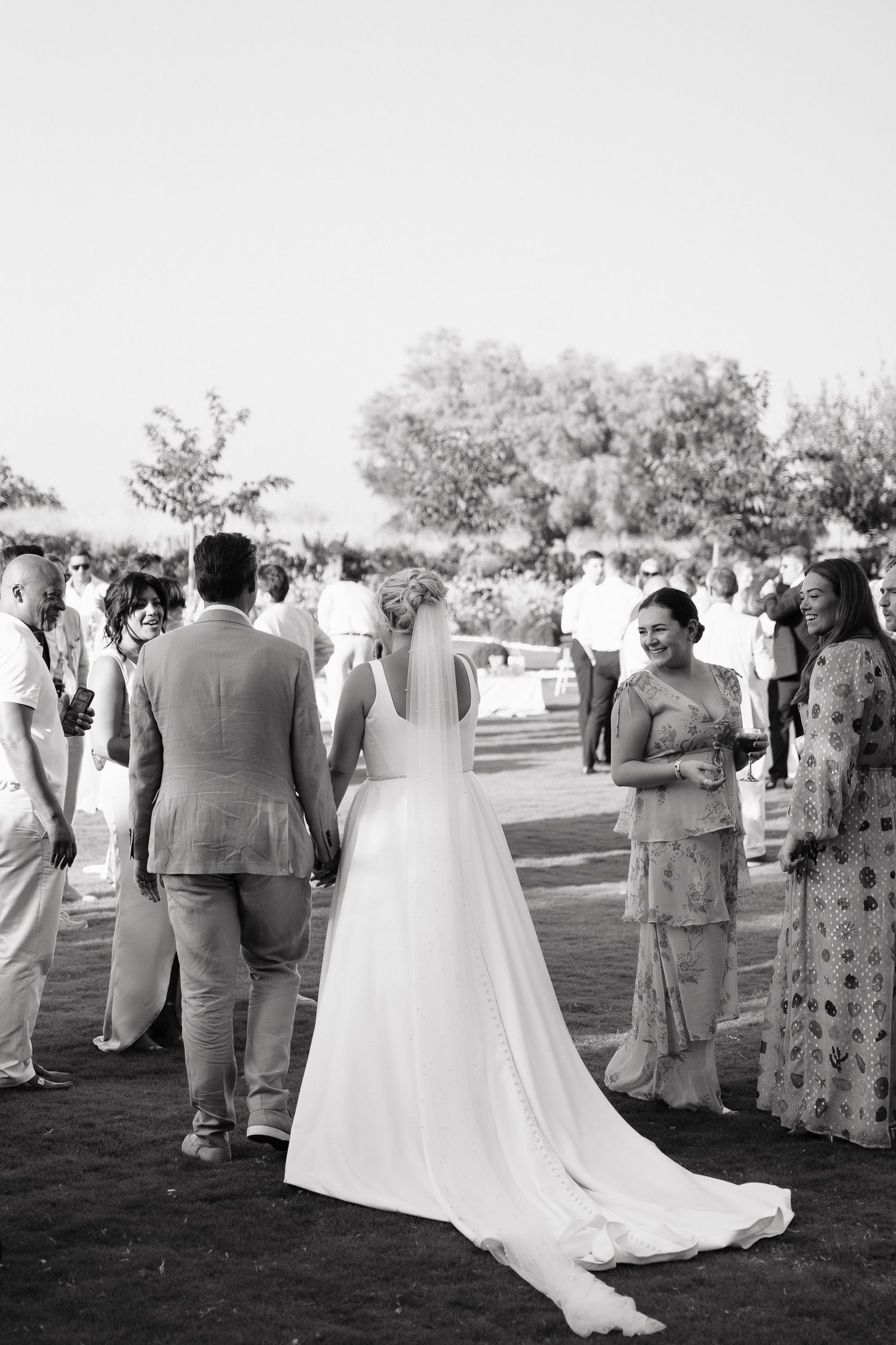 Black and white photo of an outdoor wedding reception with guests mingling on a lawn as a bride in a flowing white gown stands among them.