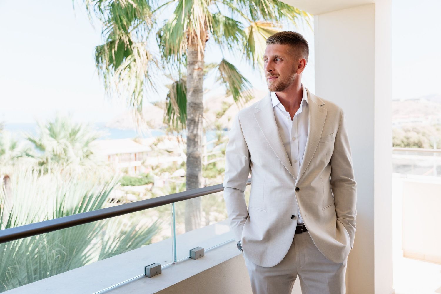 A person in a light beige suit stands on a balcony with palm trees and ocean views in the background.