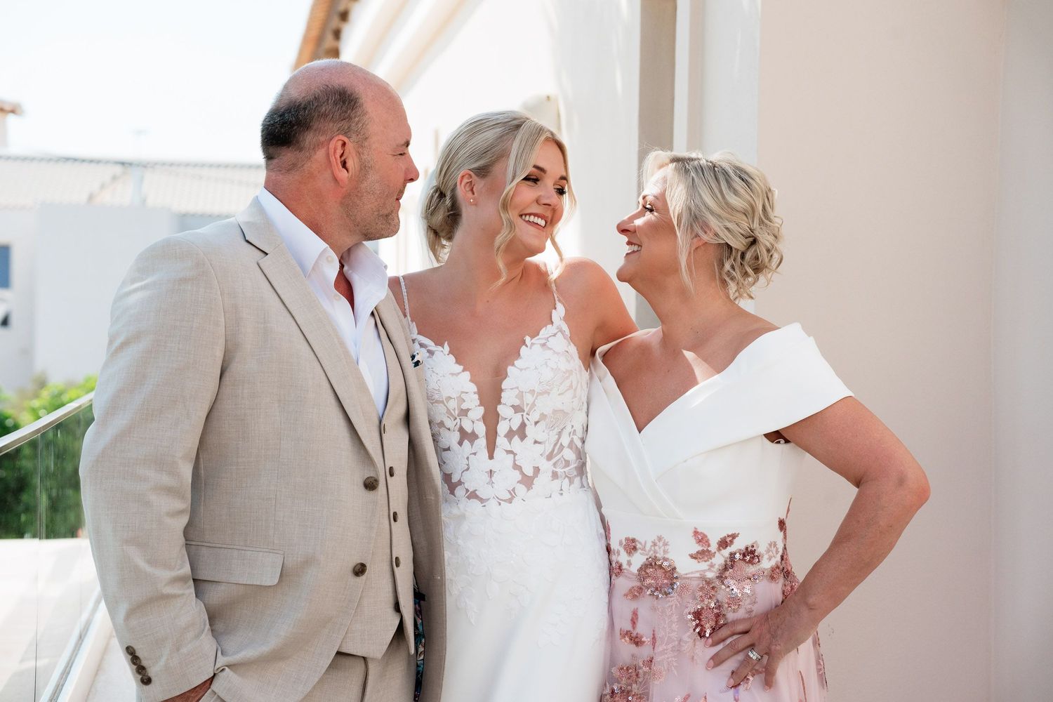 Wedding party in light summer attire laughs together during outdoor photo session at sunny venue.